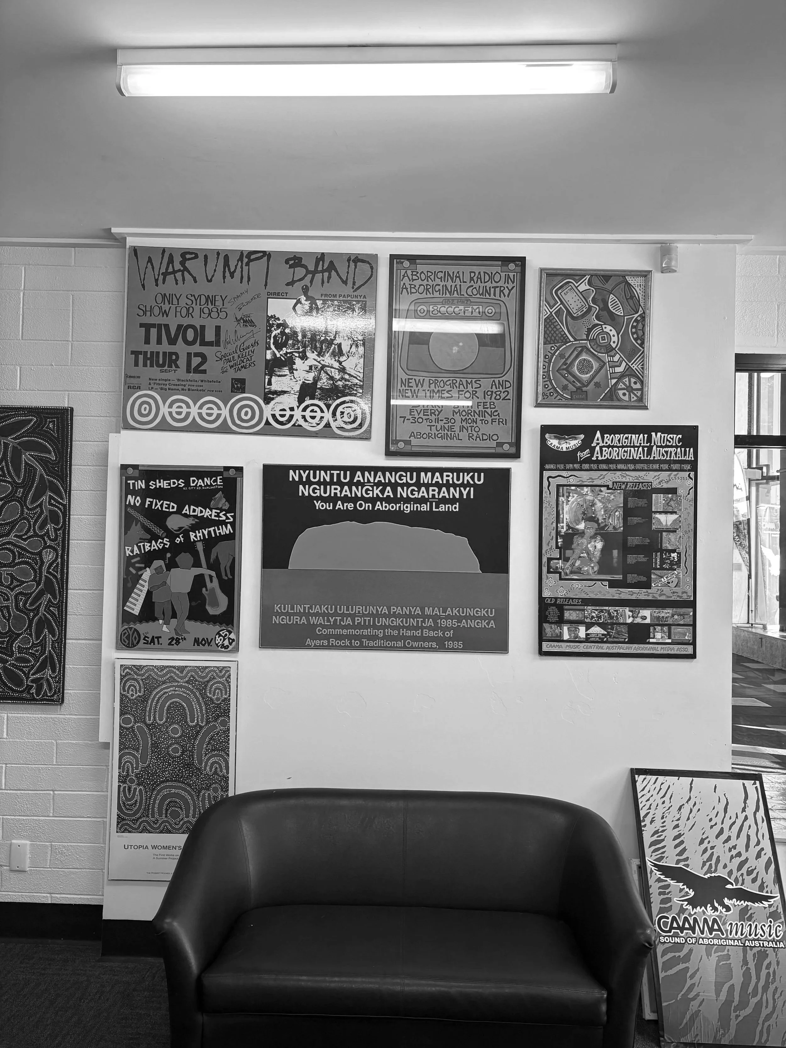 Black and white photo of a waiting area with a black chair in the center, framed posters on the wall behind it promoting Aboriginal radio, music, and cultural events, and a window on the right side.