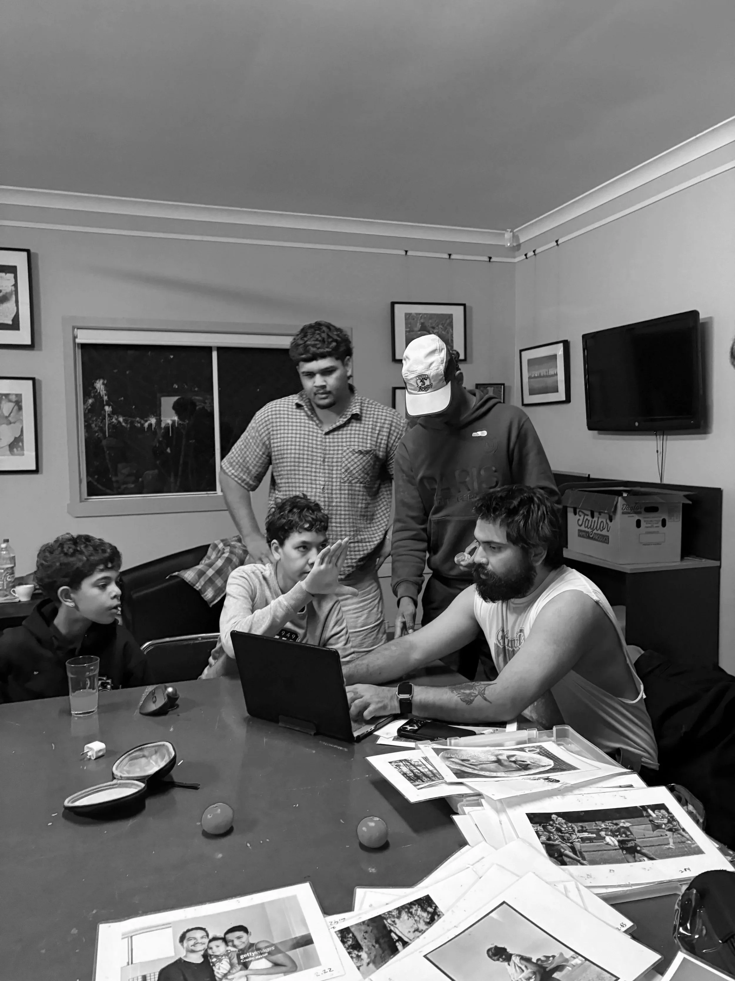 Group of young men gathered around a table with printed photographs, discussing and collaborating in a room decorated with framed pictures, a television, and boxes.