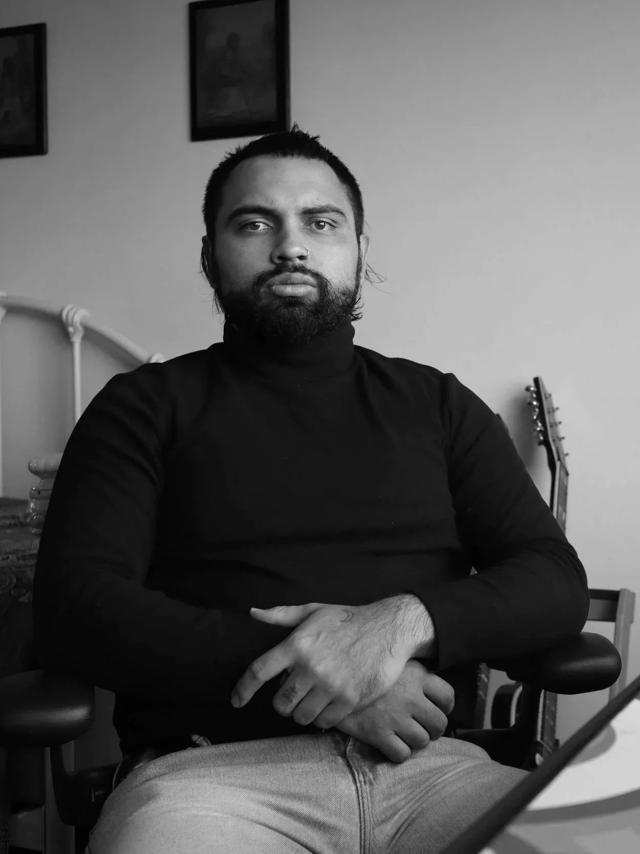 Black and white photo of a man with dark hair and a beard, sitting in a chair with arms crossed, wearing a black turtleneck shirt, in a room with pictures on the wall and a guitar in the background.