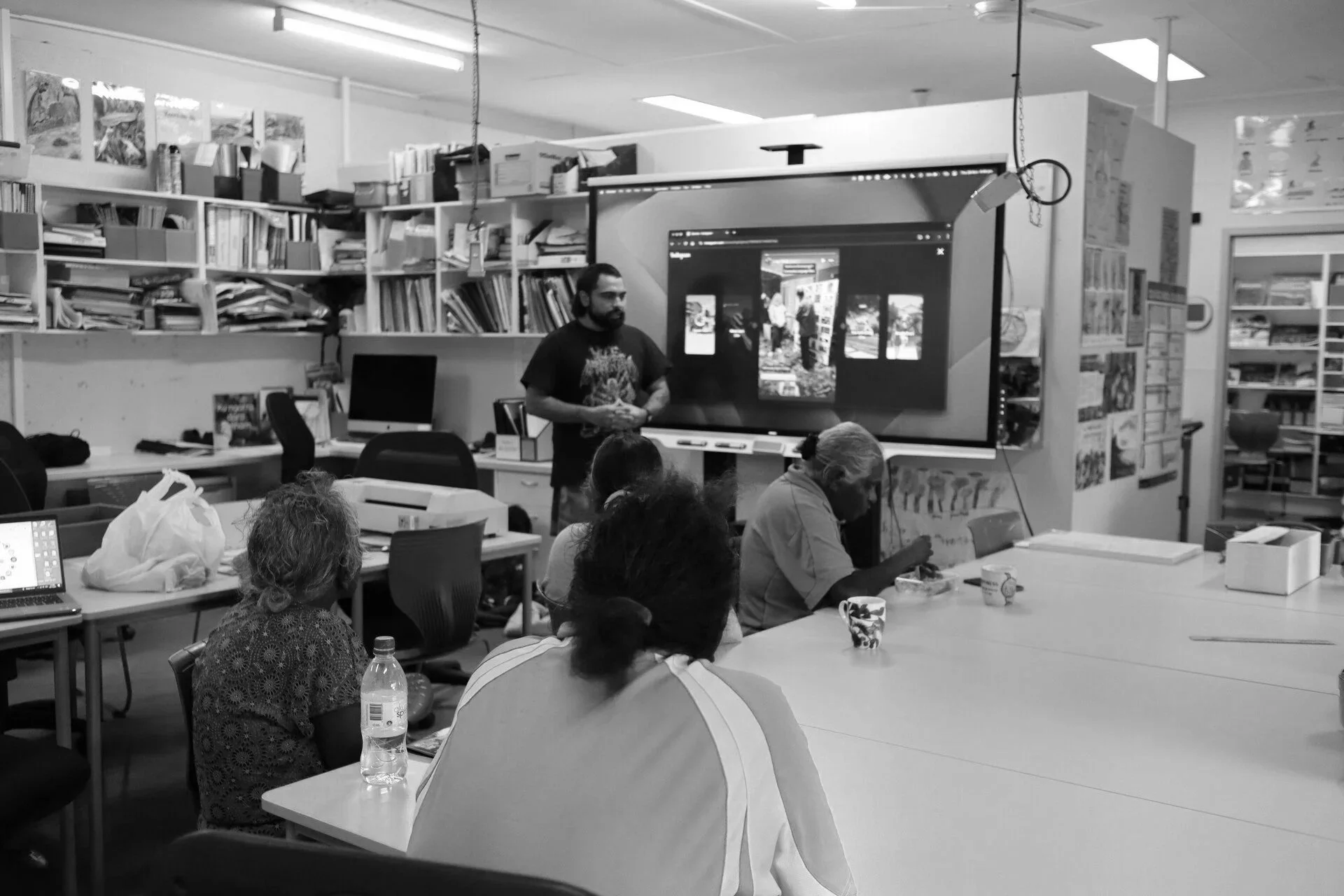 Birrugan Dunn-Velasco giving a presentation in a classroom or community center with bookshelves in the background. Several seated people are listening, some taking notes or drinking from cups.