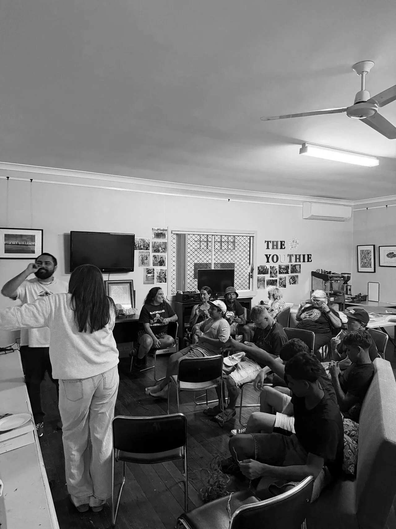 A group of people in a room with chairs, some sitting and some standing, engaging in conversation. The room has pictures on the walls, a large monitor, and a sign that reads 'The Youthie.' There are various items including a television, framed pictures, and kitchen appliances visible.