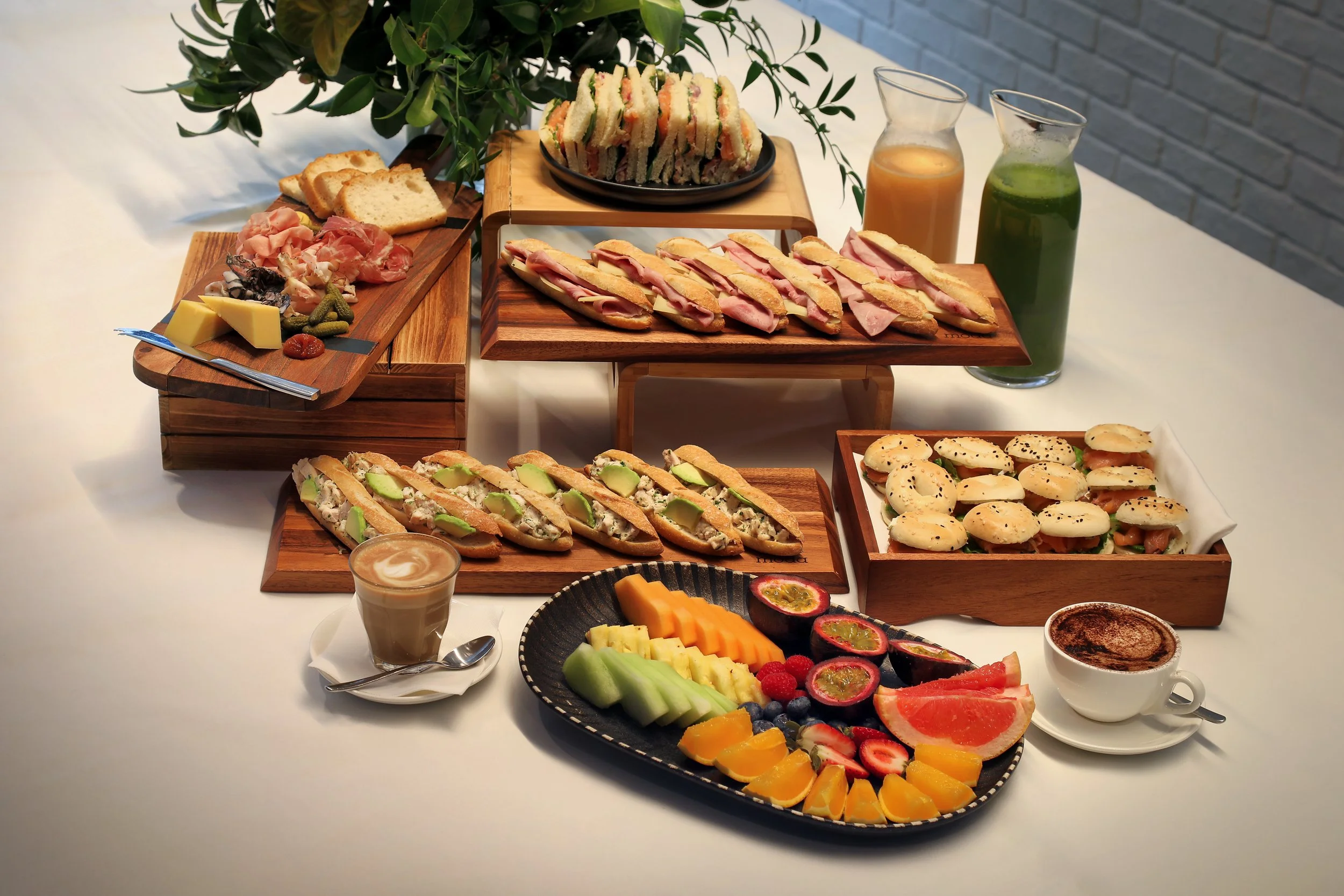 Several wooden serving platters with sandwiches, cheese, charcuterie, fruits, and beverages on a white table, with a plant in the background.