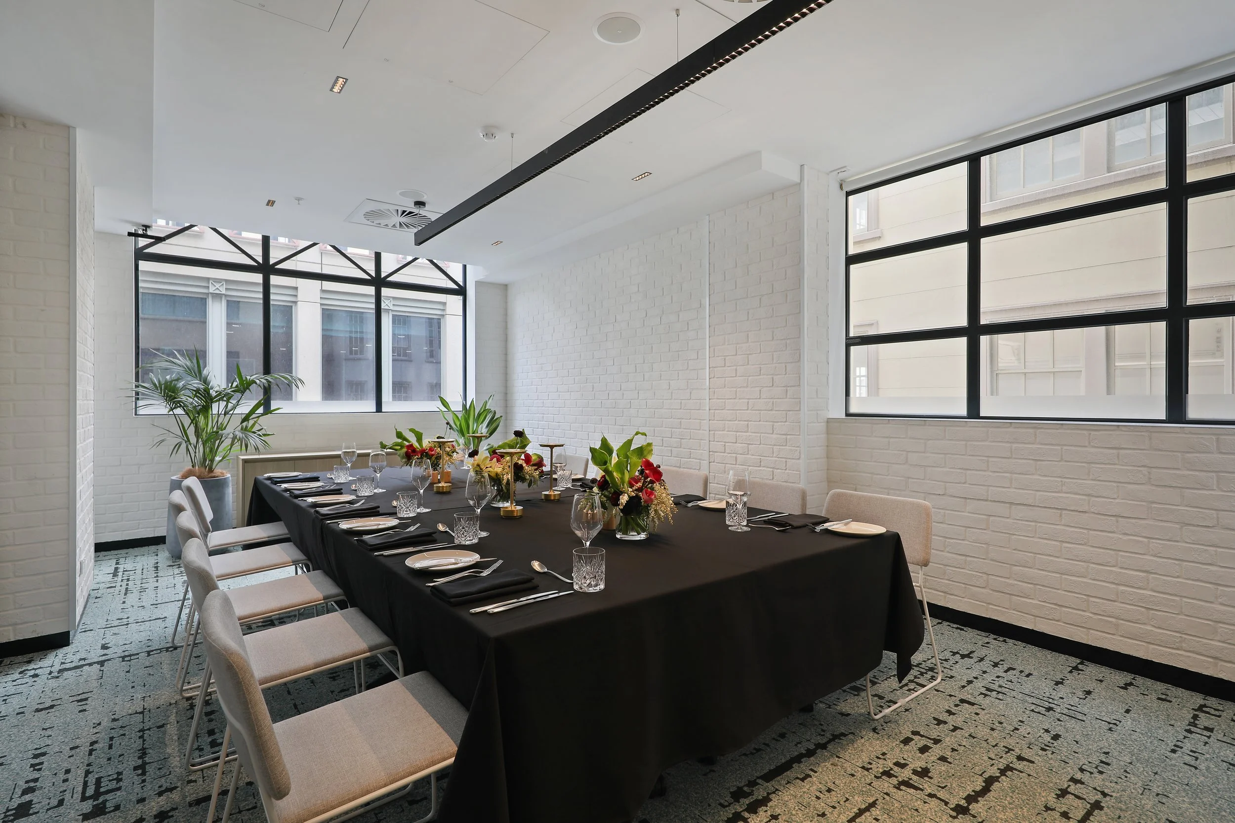 Modern dining room with a long table set for a meal, surrounded by beige chairs, decorated with flowers and tableware. Large windows with black frames allow natural light into the space, which has white brick walls and a gray patterned floor.