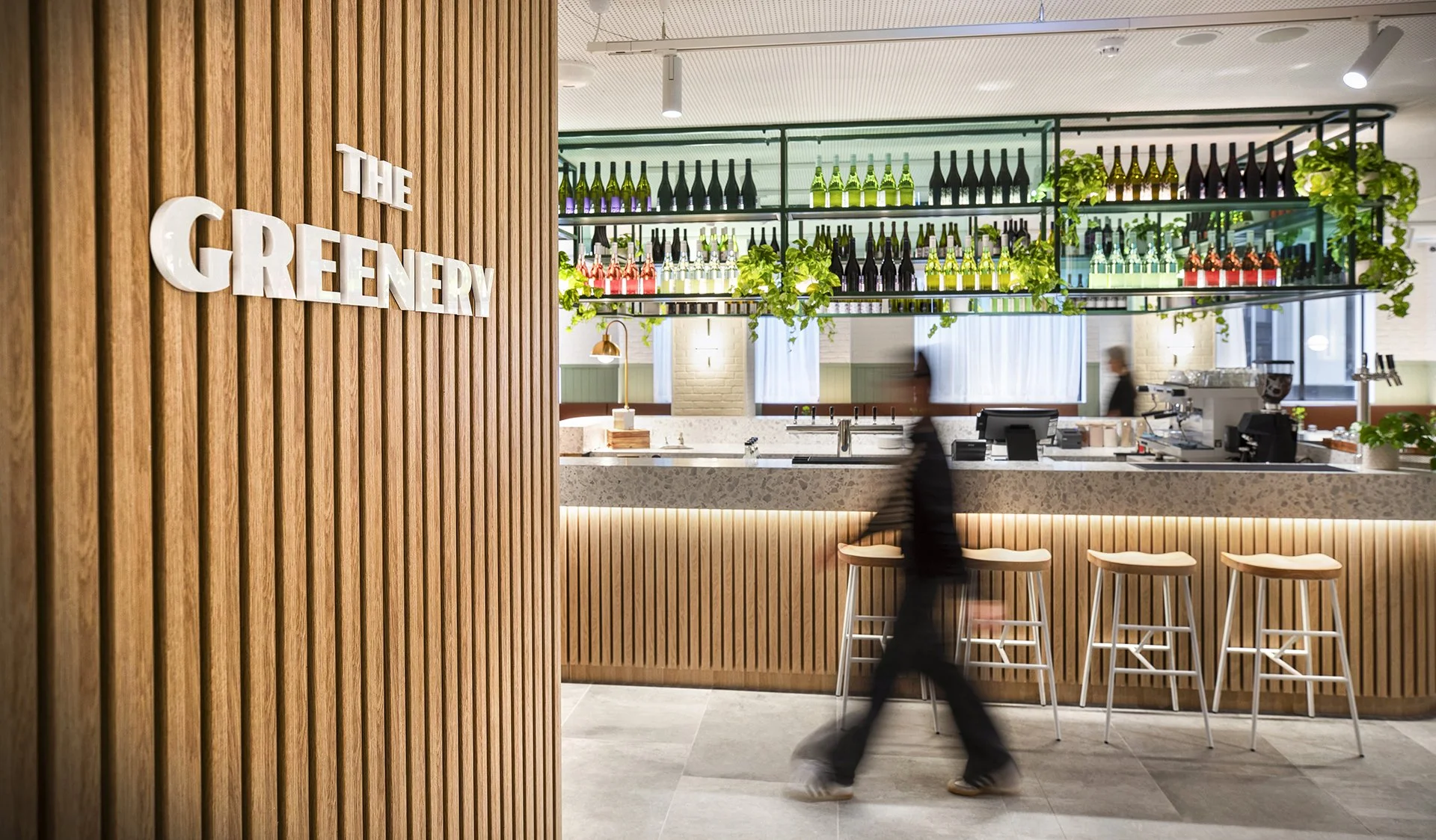 Interior of a modern restaurant or café with a wooden wall on the left that reads 'The Greenery'. Behind the bar counter, there are shelves with colorful bottles and green plants, with a barist or waiter visible in the background and three stools in front of the counter.