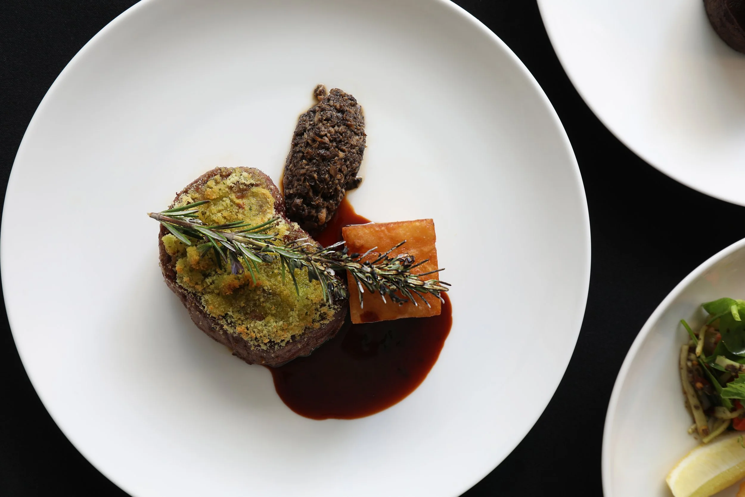 A white plate with a serving of beef Wellington garnished with a sprig of rosemary, served with a dark sauce, on a black surface.