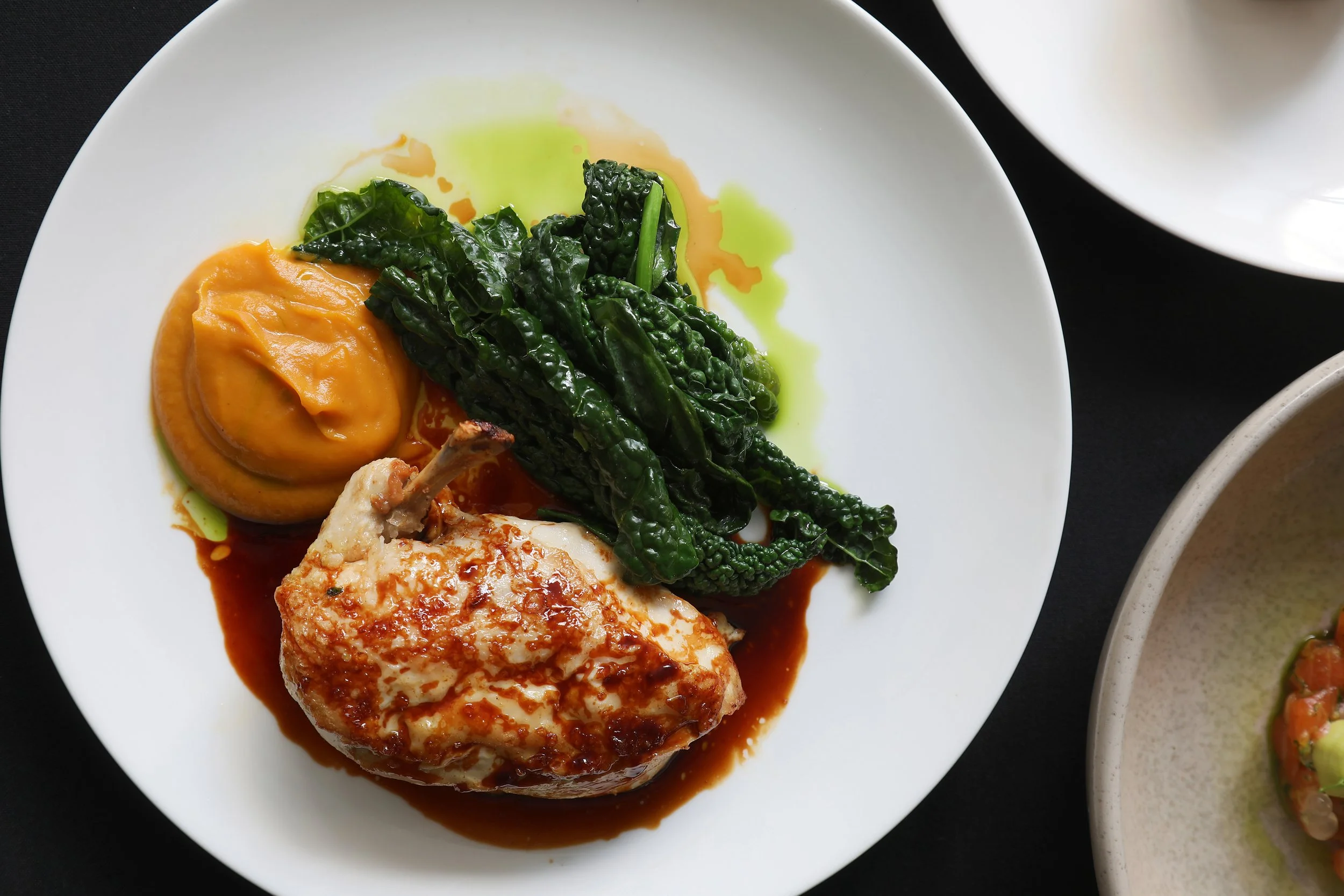 White plate containing a cooked chicken breast topped with a brown sauce, served with wilted greens and orange mashed sweet potatoes, on a black surface.