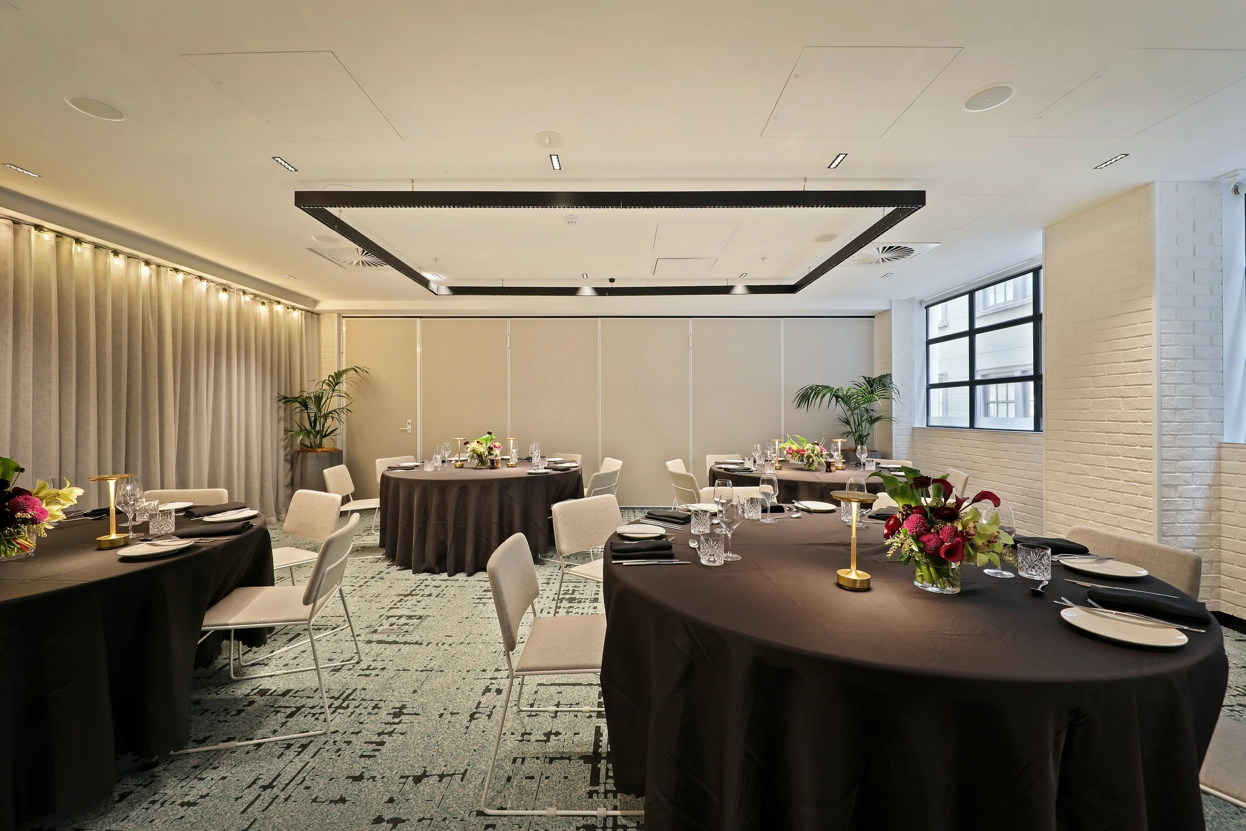 Elegant banquet room with round tables covered in black tablecloths, each set with glassware, plates, and black napkins. Centerpieces feature colorful flowers and gold candlesticks. The room has large windows, white brick walls, and beige curtains, w