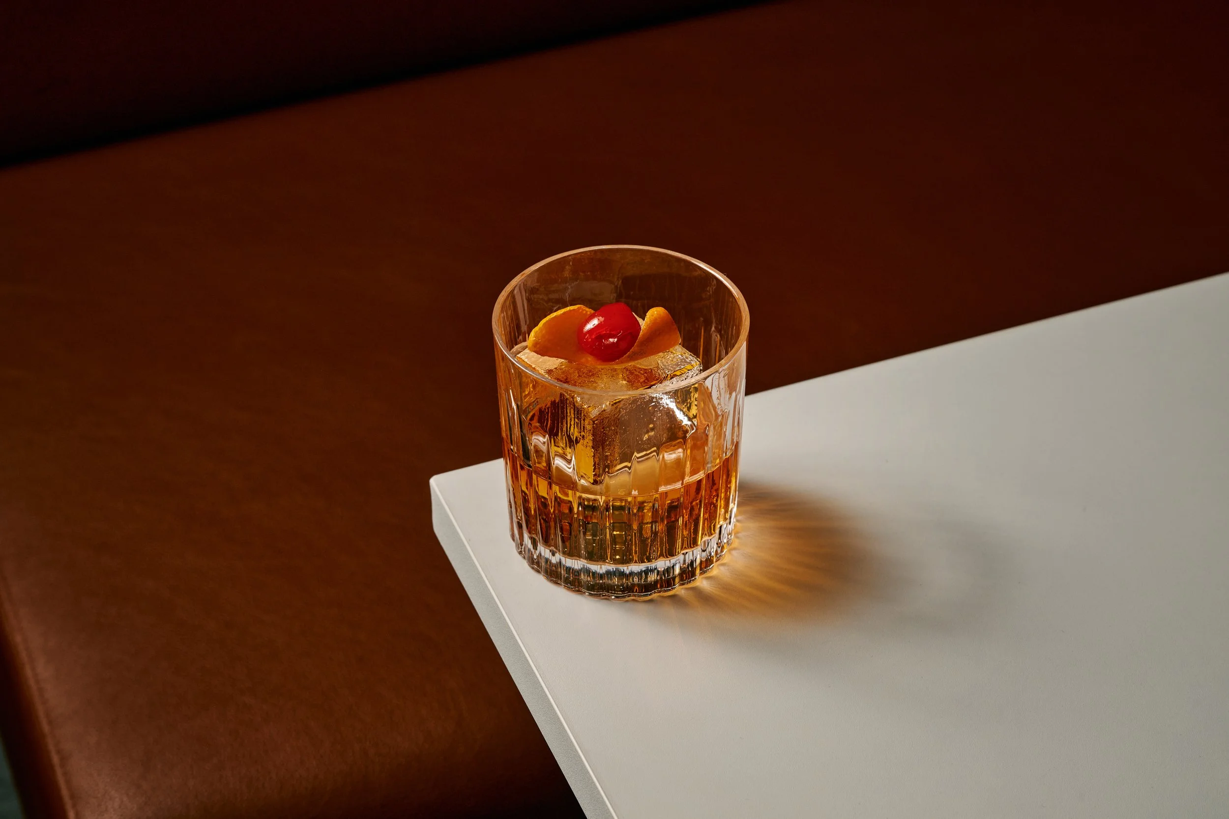 A glass of amber-colored beverage with ice, garnished with orange peel and maraschino cherry, on a white surface.