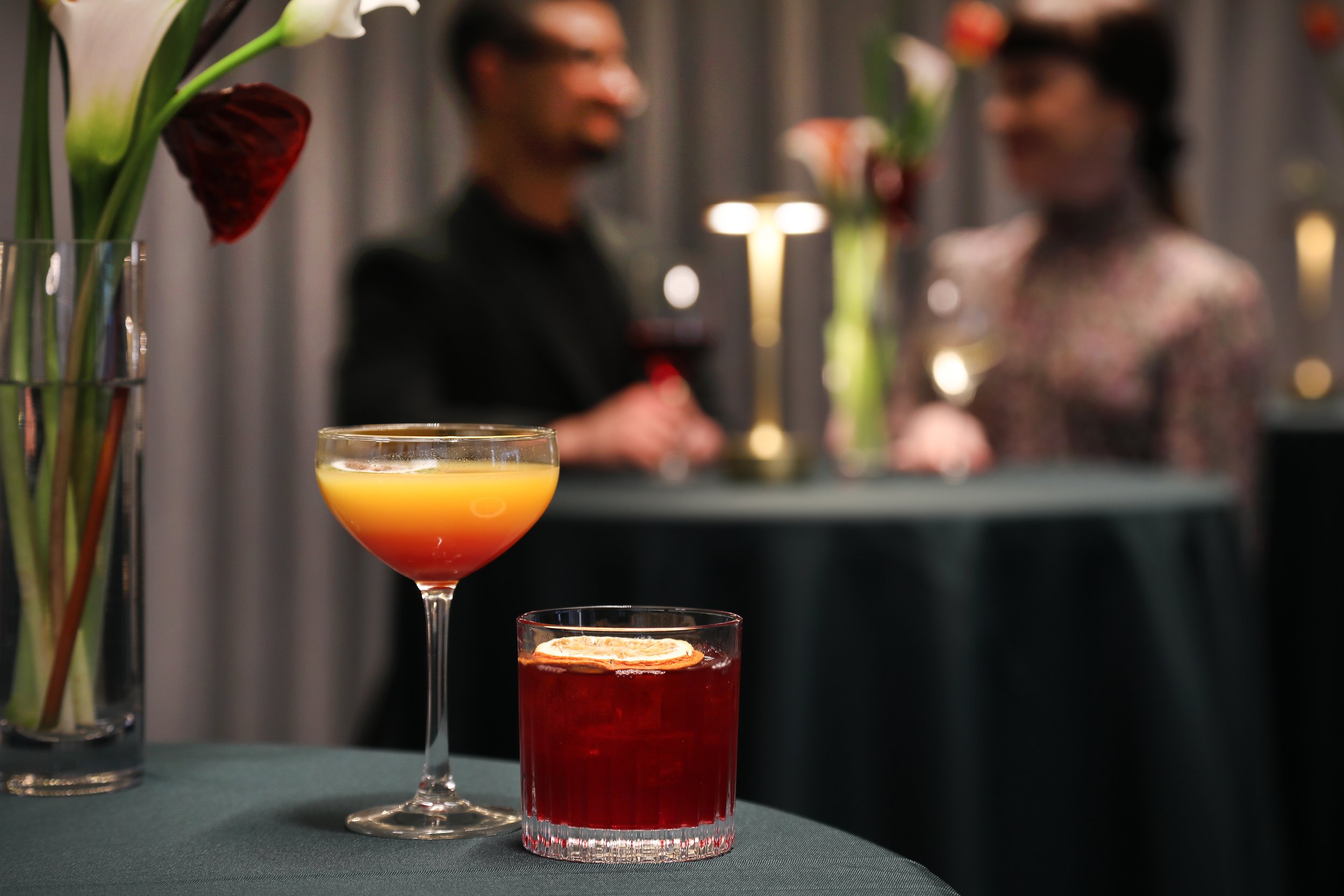 Close-up of a cocktail with yellow and orange layers in a coupe glass. In the background, two people are blurred, smiling and holding drinks, with a decor of flowers and dim lighting.