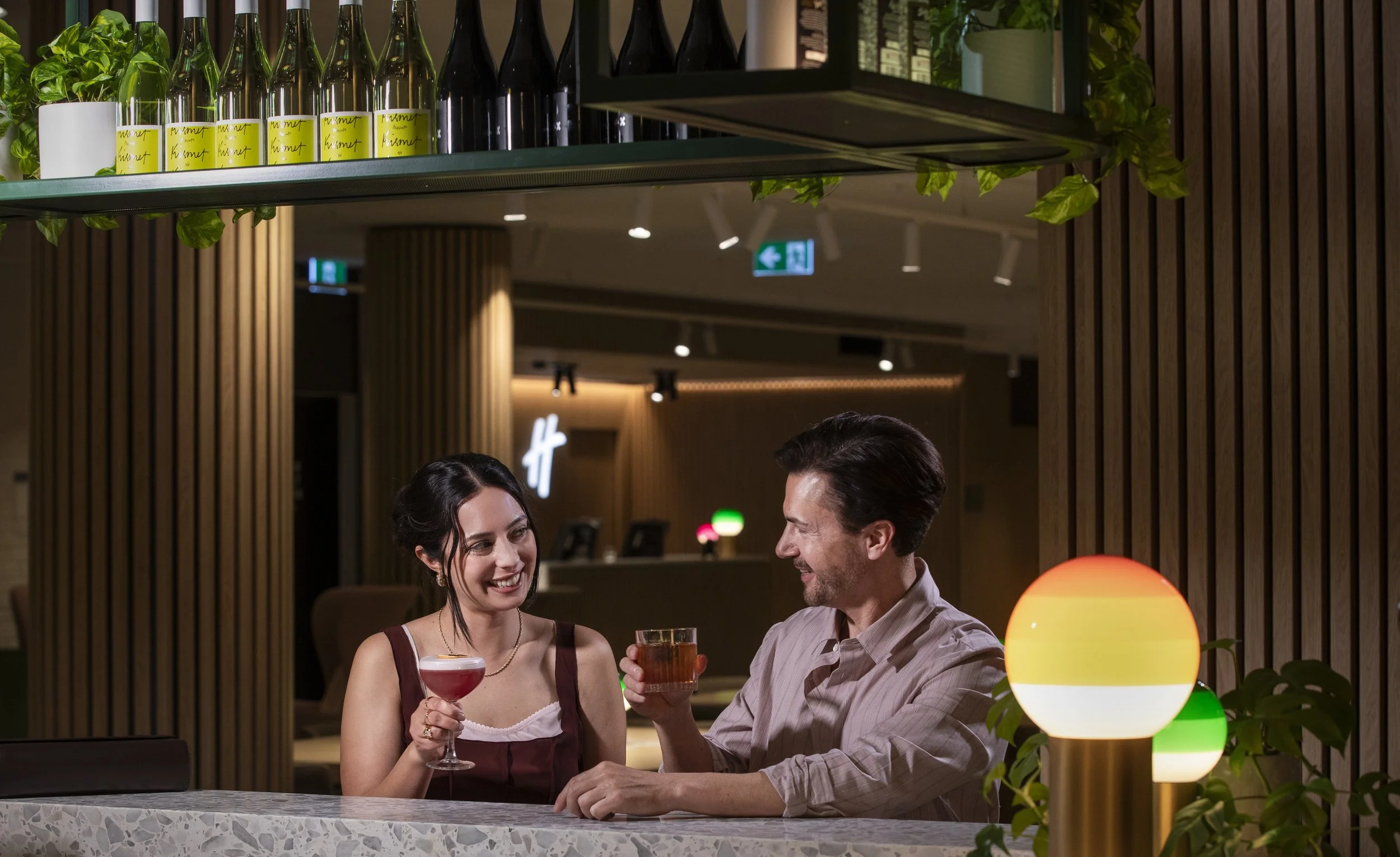 A man and a woman sitting together at a bar, smiling and holding glasses of drinks, with warm ambient lighting and wooden paneling behind them.