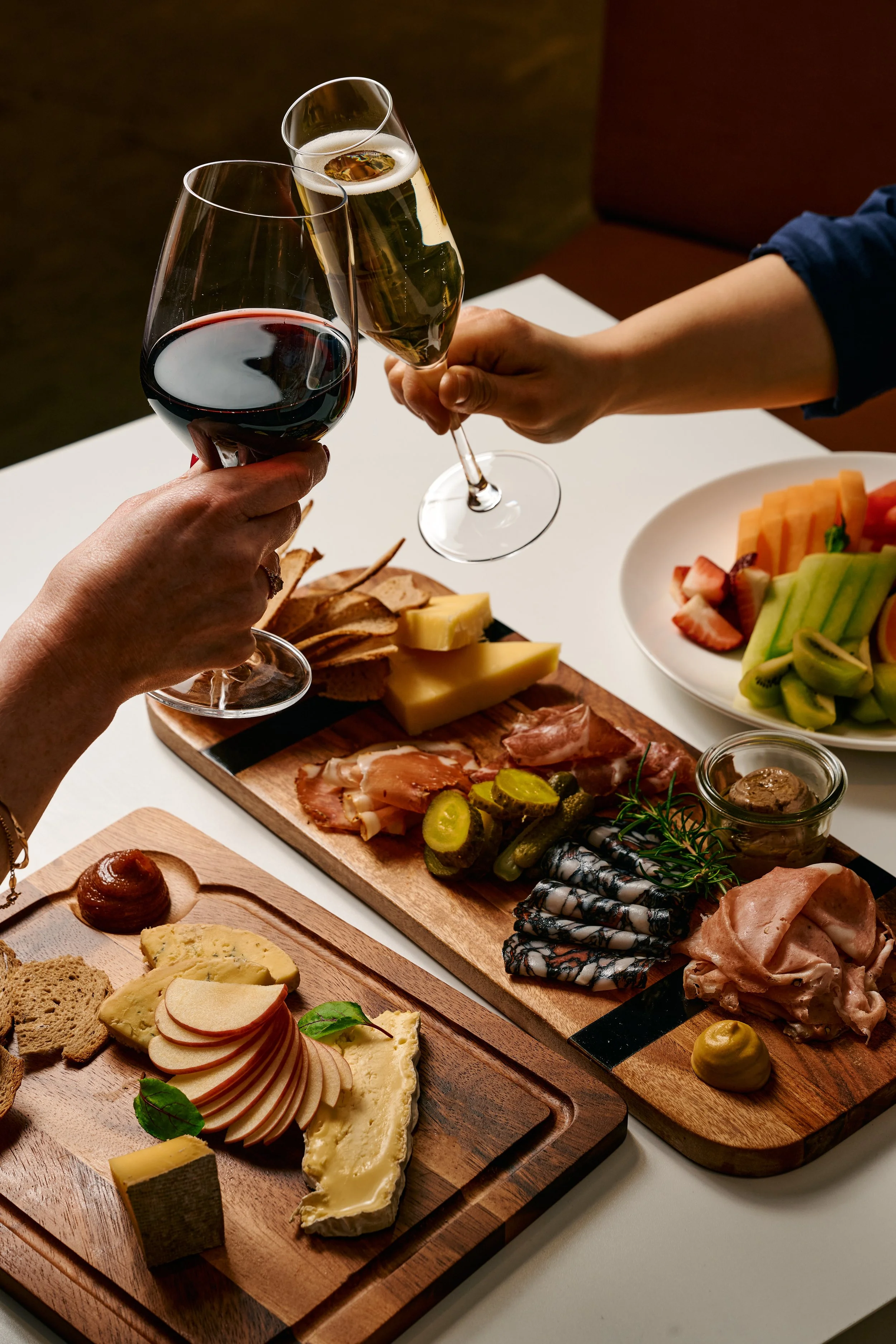 Two people clinking glasses of wine, one red and one white, over a table with assorted cheeses, charcuterie, fruit, and crackers.