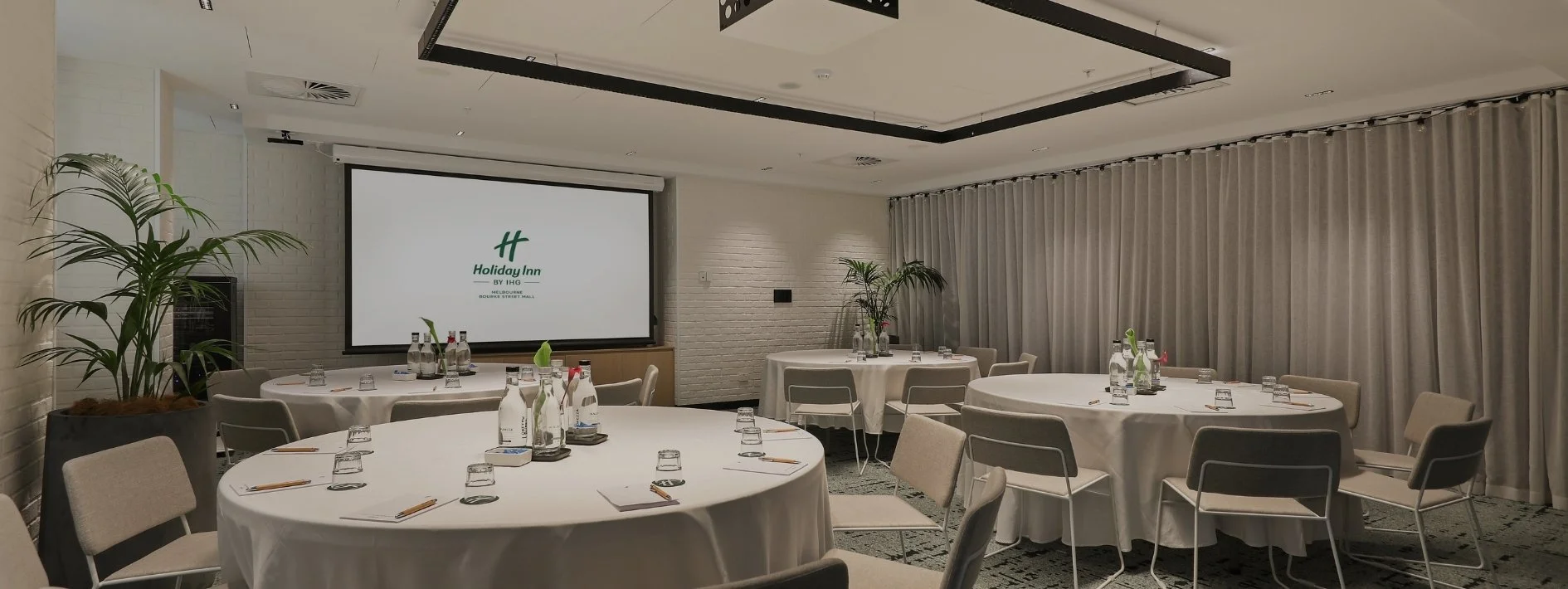 A conference room set up with round tables covered in white tablecloths, each with bottled water, glasses, and notepads. There are chairs around each table, potted plants for decoration, a large screen displaying a Holiday Inn logo, and beige curtains along the windows.