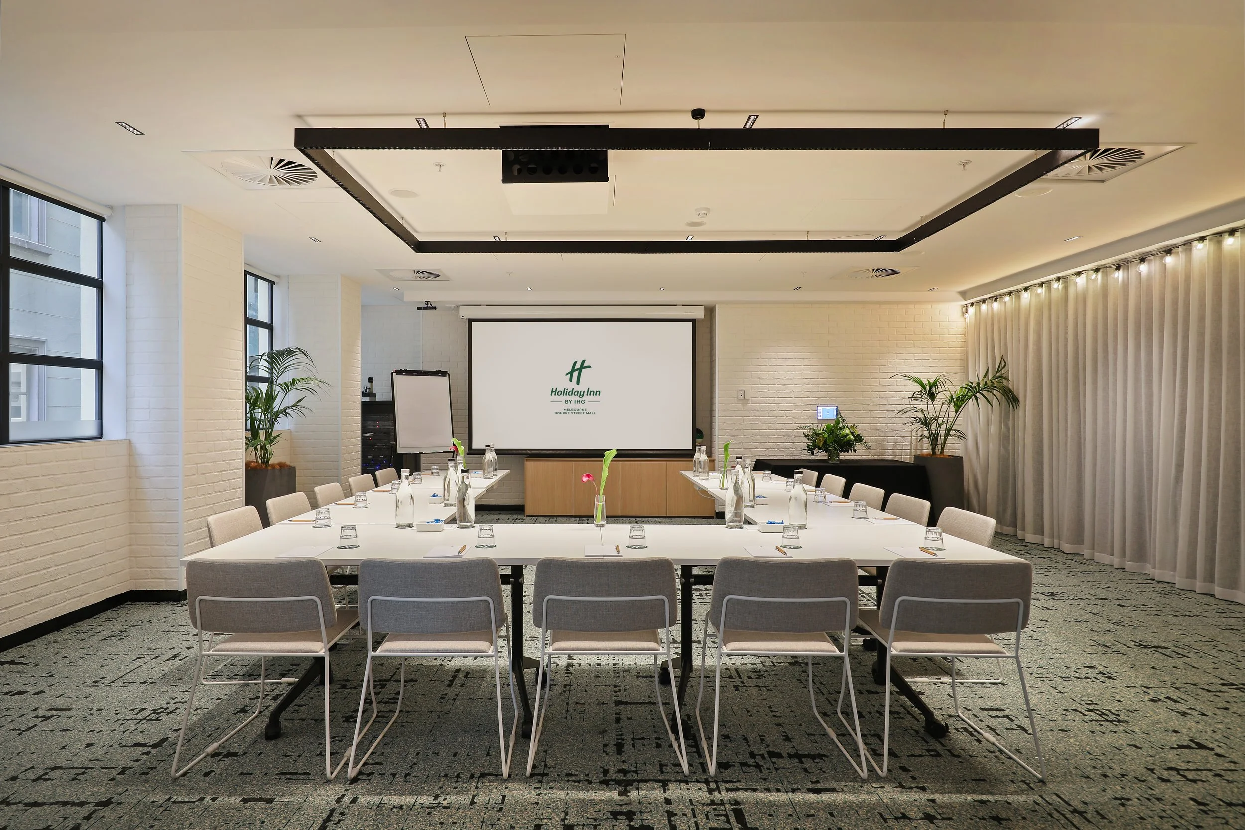 Conference room set up for a meeting with tables, chairs, water bottles, and notebooks. A large screen displays the Holiday Inn logo, and there are windows, plants, and curtains in the room.