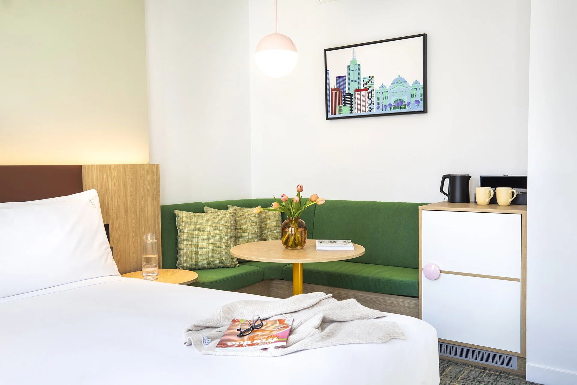 A hotel room with a made bed, a nightstand with a glass of water, and sunglasses on a magazine. The room has a green corner sofa with plaid cushions, a wooden table with a vase of tulips, a framed cityscape picture on the wall, and a small kitchenette area with a coffee pot, mugs, and a black box.