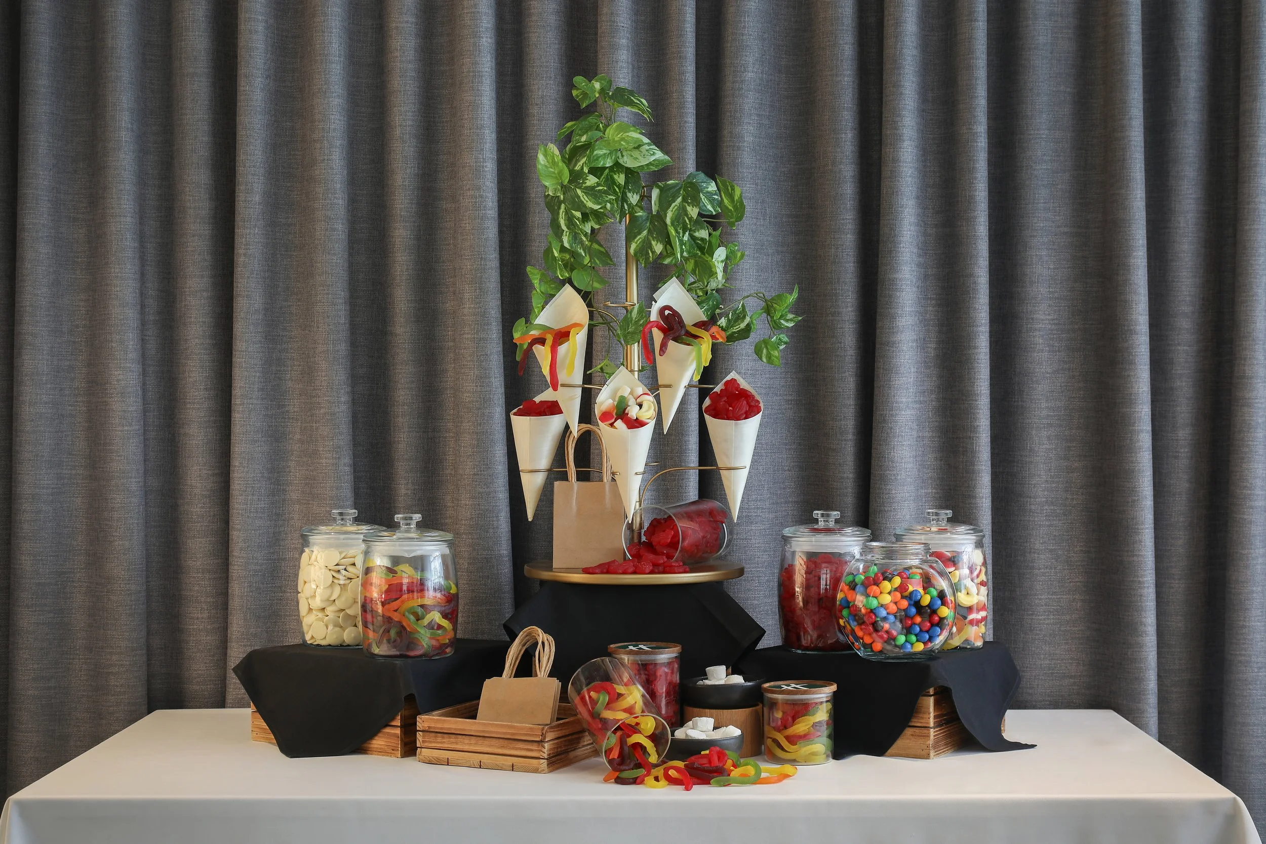 Candy display with jars of gummy bears, gummy worms, and M&Ms, with a decorative plant and a cake on a table covered with black and white cloths, set against a dark gray curtain background.
