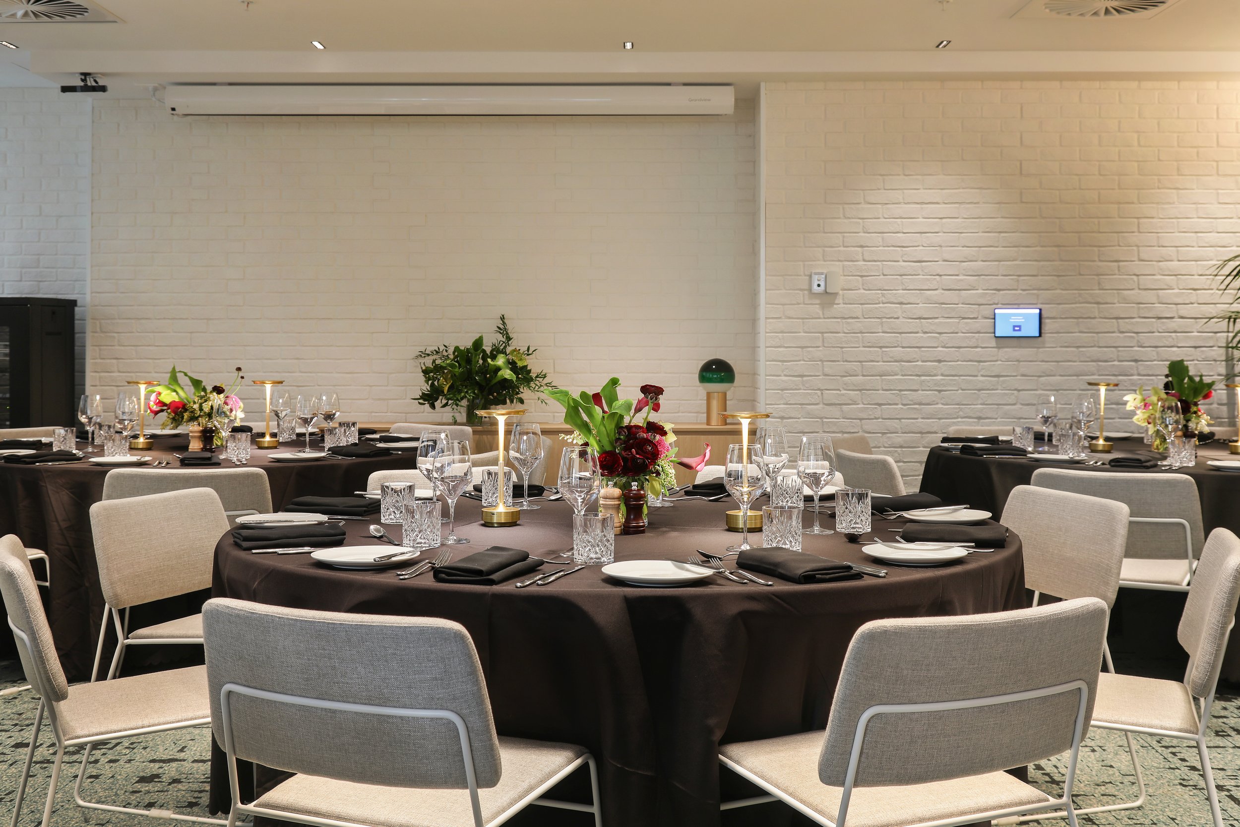 Elegant dining setup with round tables, black tablecloths, white napkins, and floral centerpieces in a modern space with white brick walls.