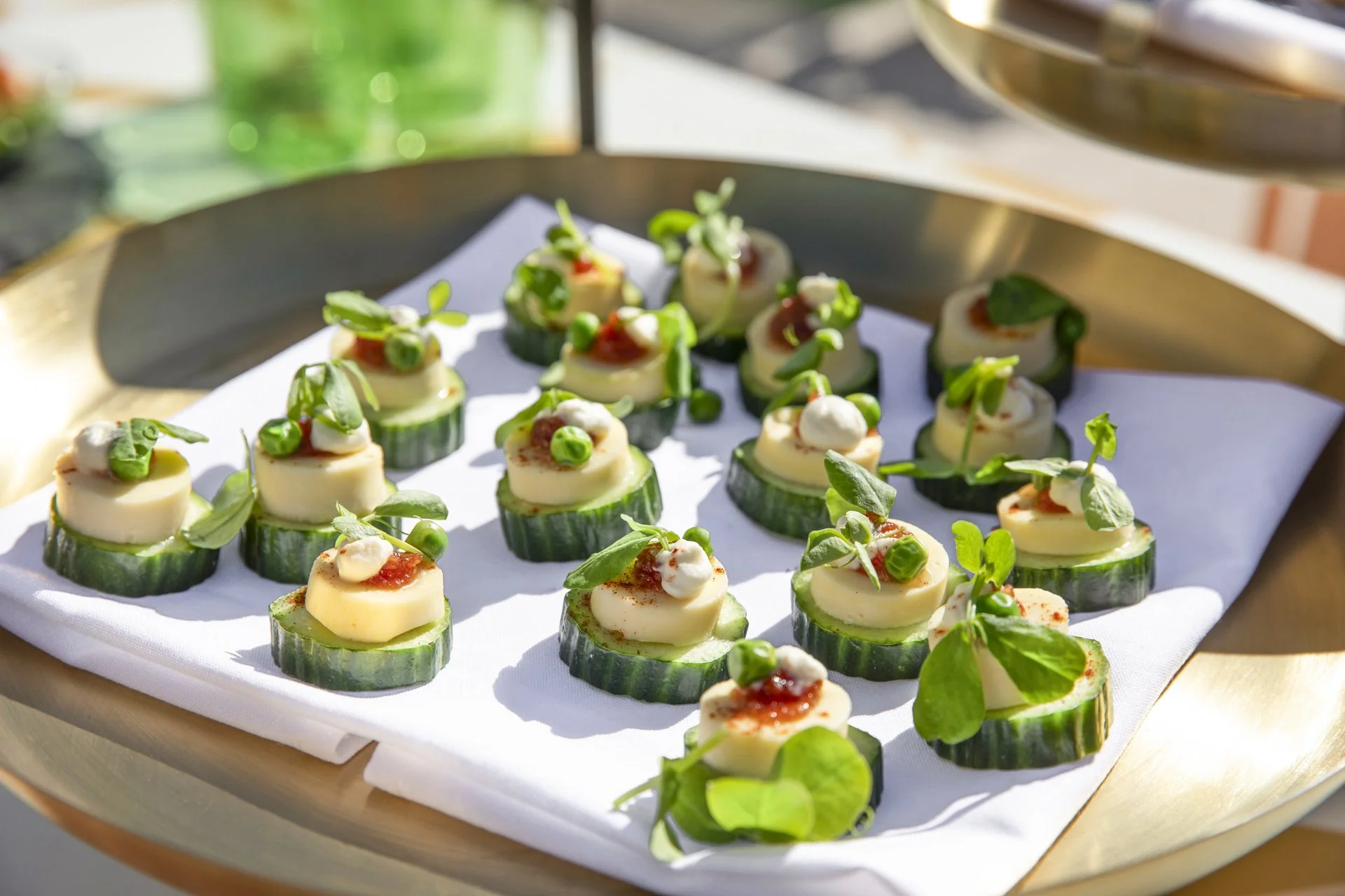 Cucumber slices topped with cheese, cherry tomato, green pea, and microgreens on a white napkin lined tray.