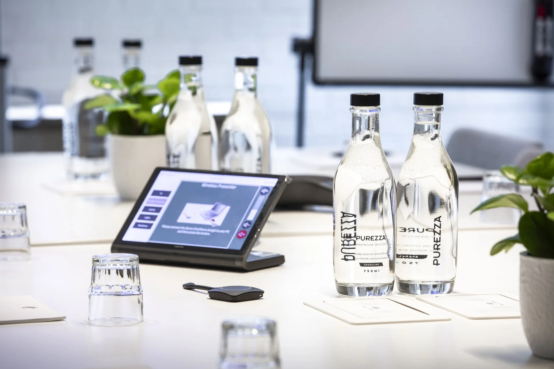 A conference room table setup with water bottles labeled PUREZZA, a tablet displaying a presentation, empty glasses, small potted plants, and a large monitor screen in the background.