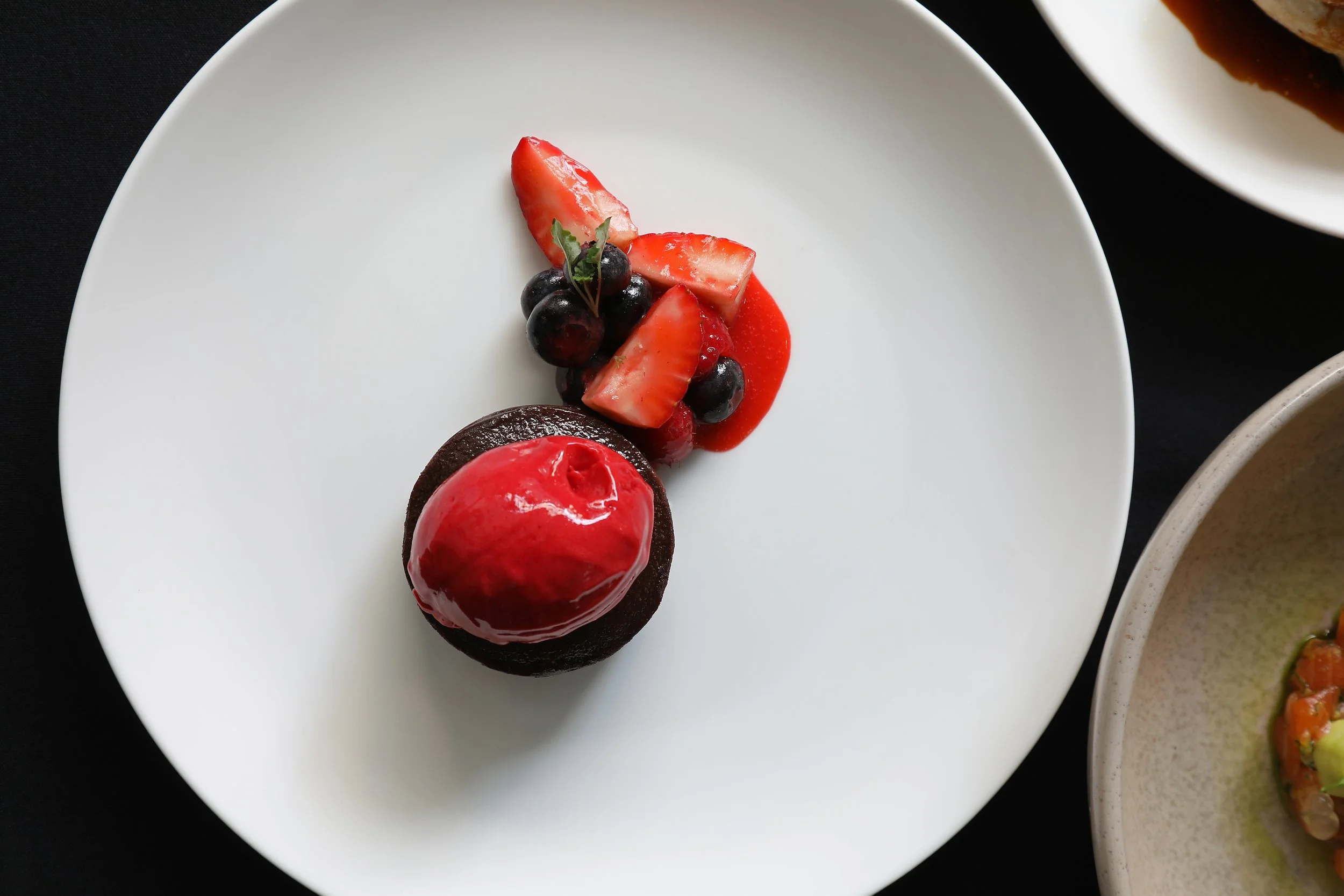 A white plate with a dessert featuring a glossy red dome, decorated with a small piece of greenery. The plate also has a candy or biscuit topped with a glossy red glaze, surrounded by slices of strawberries and blueberries, with a red sauce drizzled 
