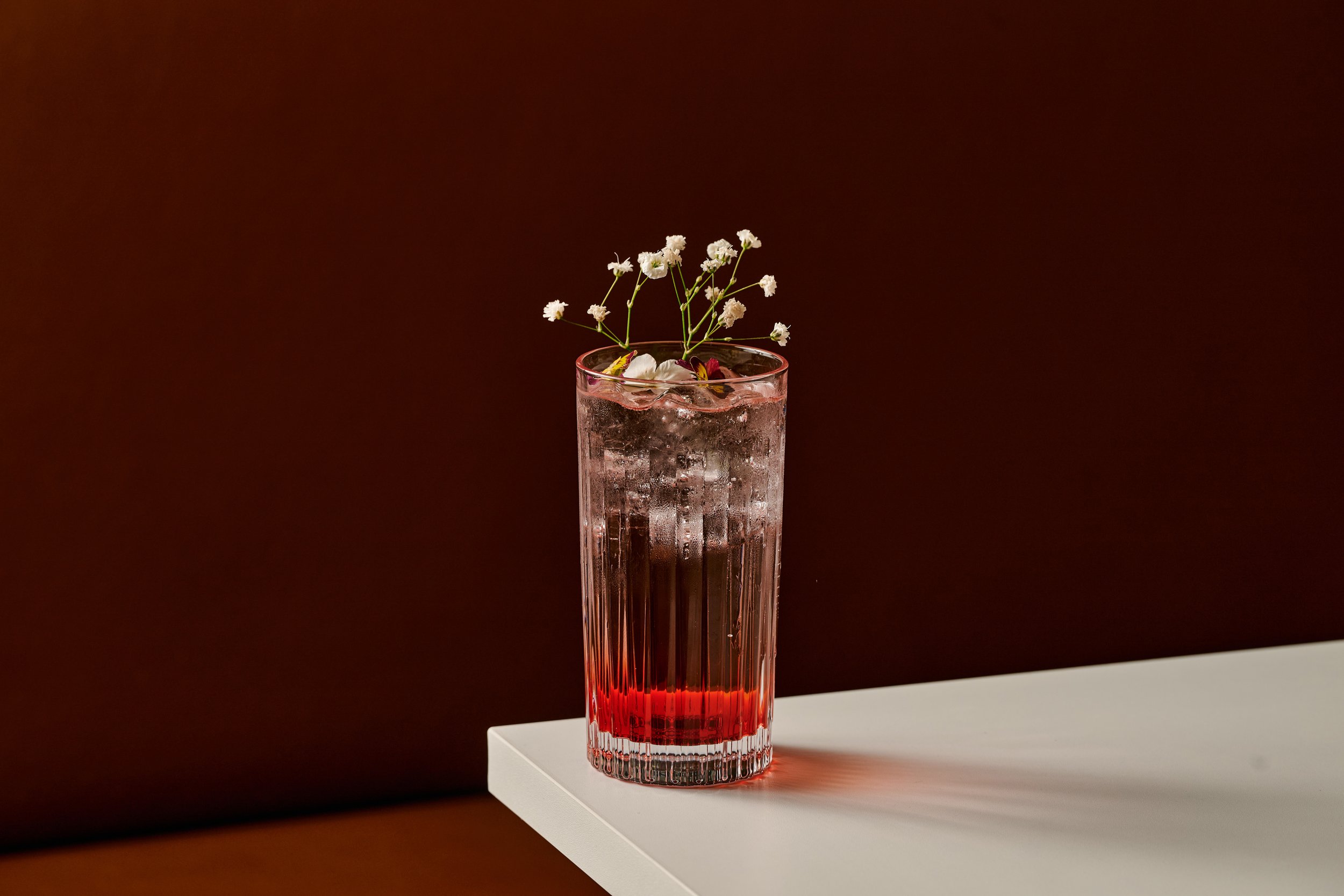 A tall, ribbed glass filled with a pink-colored beverage, garnished with white baby's breath flowers, resting on a white surface against a dark background.