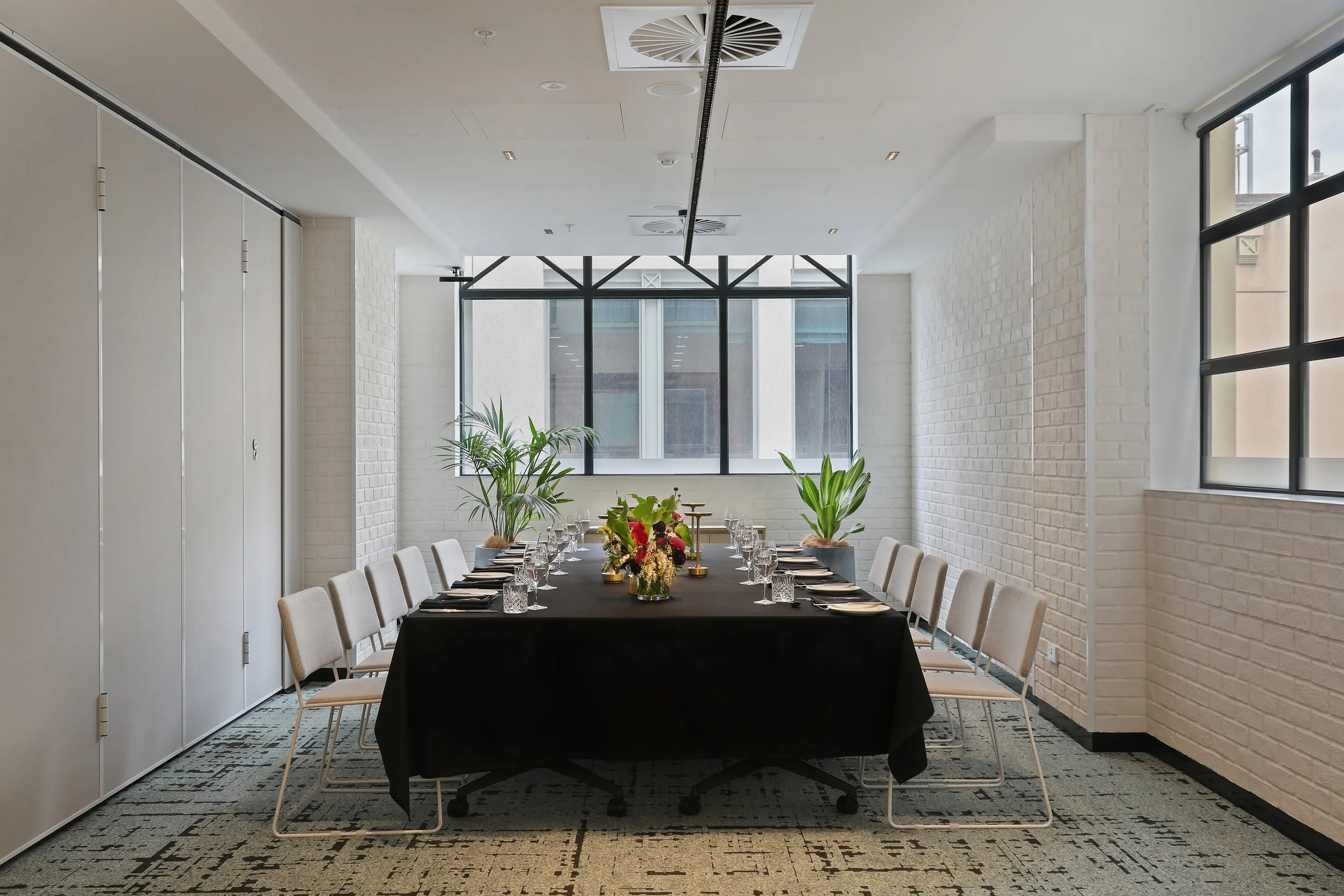 A modern conference room with a long black table set for a meeting, surrounded by beige chairs, with large windows and decorative plants, in a bright space with white brick walls.