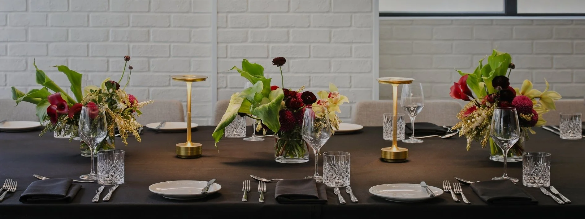 Elegant table setting with floral centerpieces, wine glasses, plates, and cutlery, set against a white brick wall.