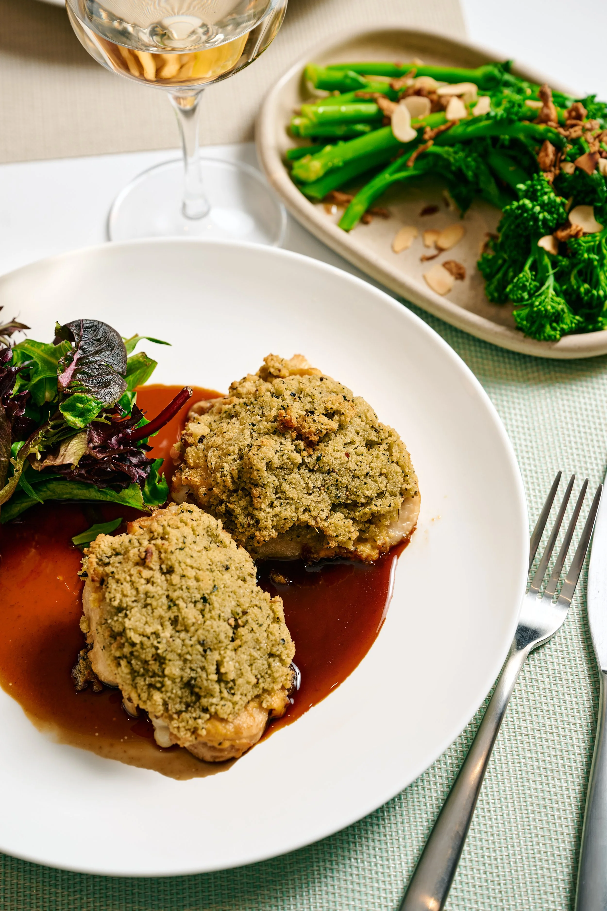 A plate with three pieces of baked food topped with a crumbly coating, served with a brown gravy and a side salad of mixed greens. In the background, a dish of cooked broccolini sprinkled with sliced almonds is visible. A glass of white wine is also 
