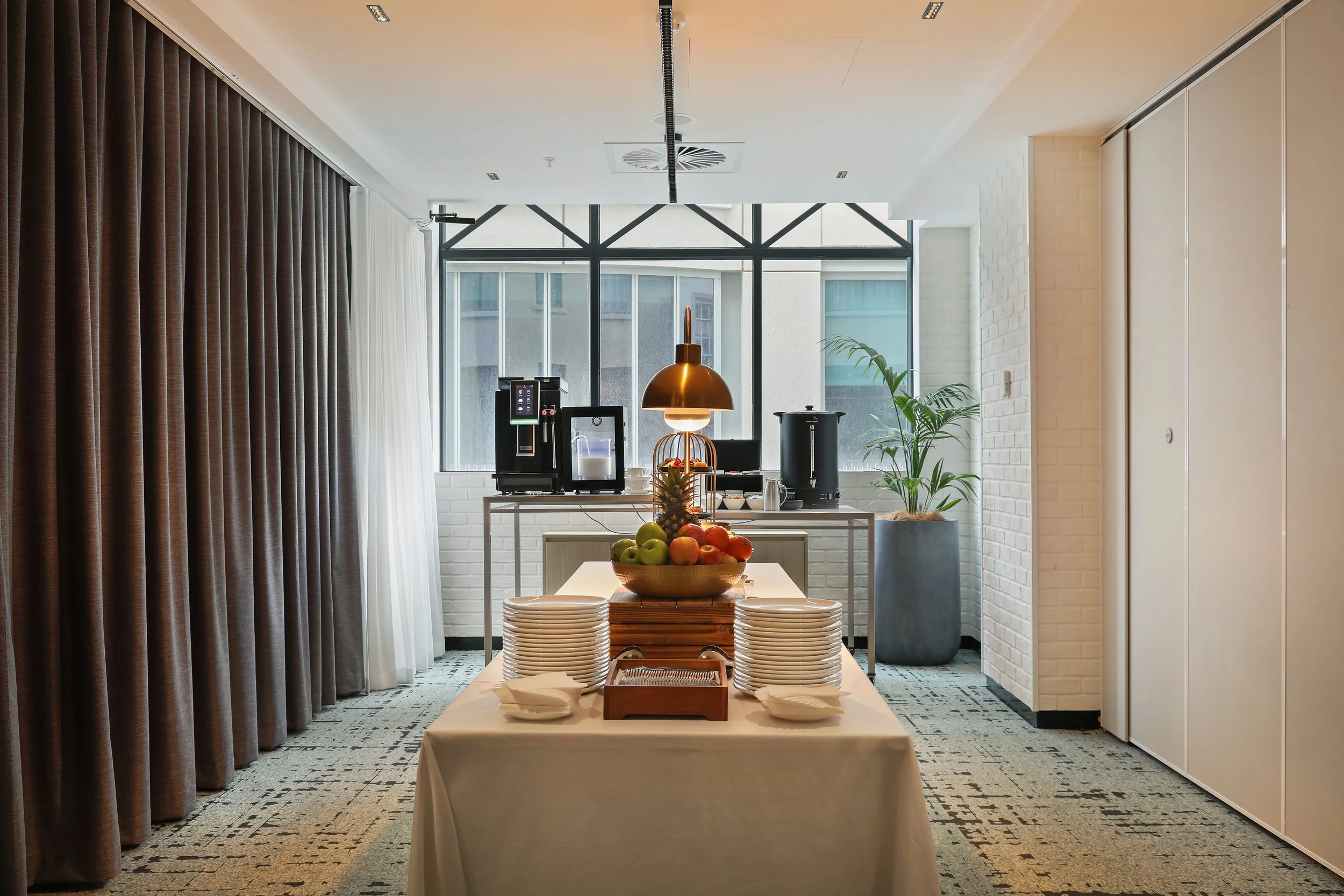 Interior of a modern dining area with a table set with a bowl of assorted fruits and plates, facing a window with black framing, with a coffee station and a plant in a gray pot in the background.