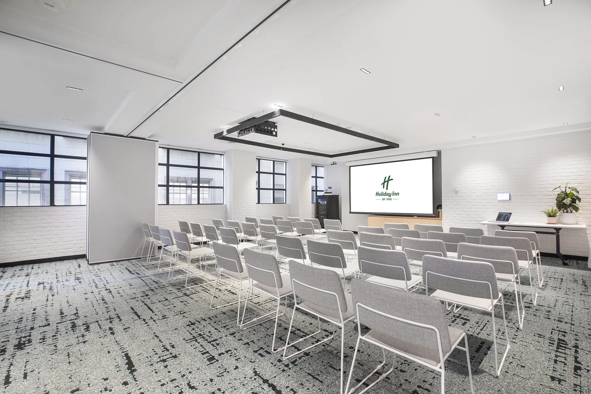 Empty conference room with chairs facing a large screen displaying the Holiday Inn logo, windows on the left, and a table with plants on the right.