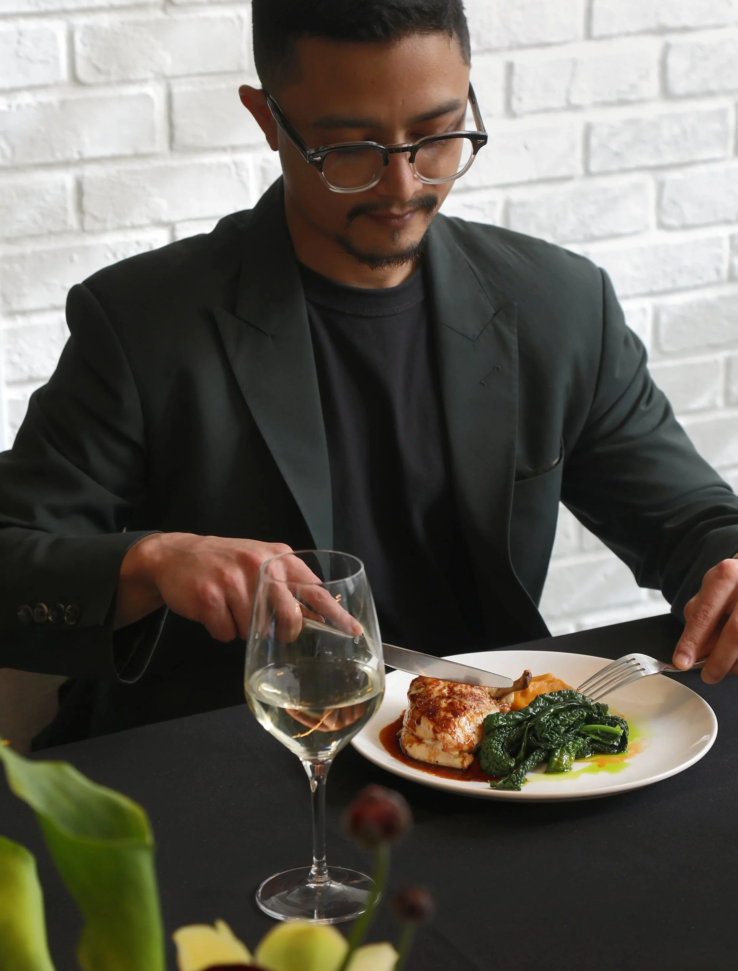 A man with black hair and glasses wearing a black suit is sitting at a table, cutting food on a white plate that has greens and a piece of meat. There is a glass of white wine in the foreground and some yellow and green flowers in the bottom left corner.
