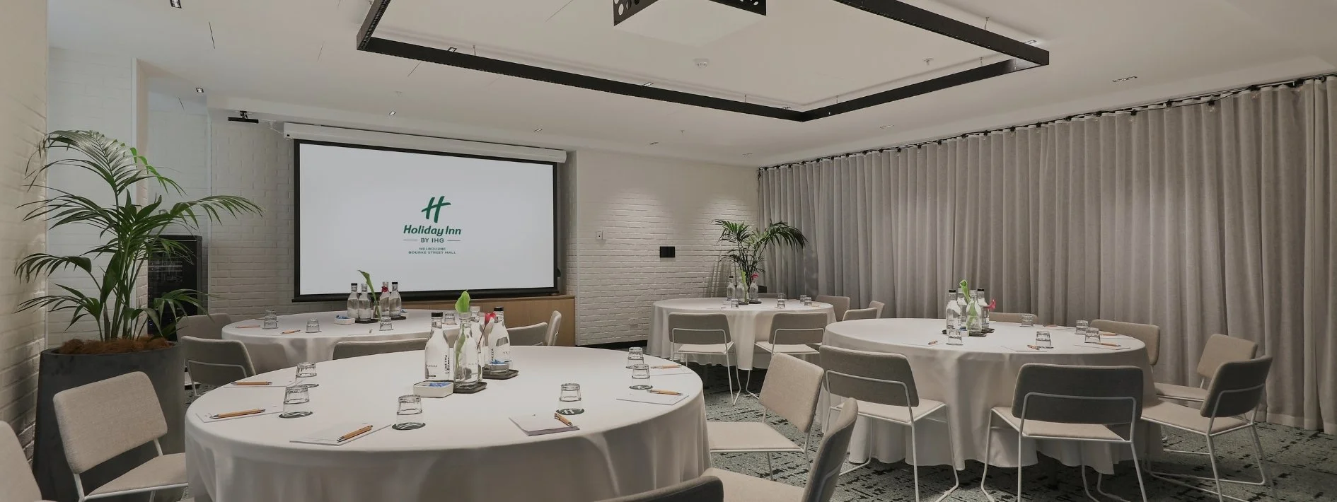 A conference room set up with round tables covered in white tablecloths, each with bottled water, glasses, and notepads. There are chairs around each table, potted plants for decoration, a large screen displaying a Holiday Inn logo, and beige curtains along the windows.