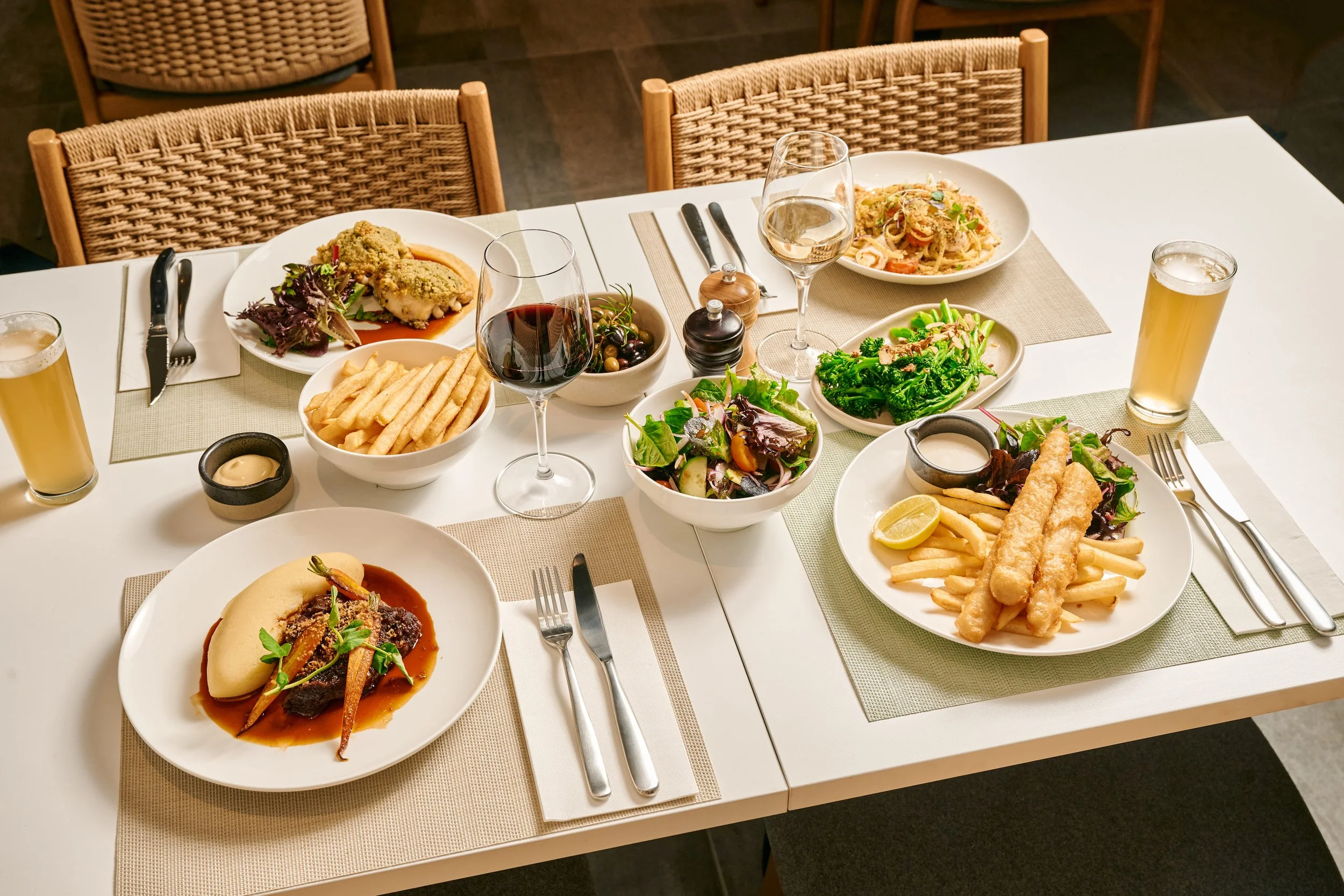 A table set for a meal with various dishes, including fried fish with fries, pasta, a salad, a burger, and drinks like wine and beer, in a restaurant setting with woven chairs.