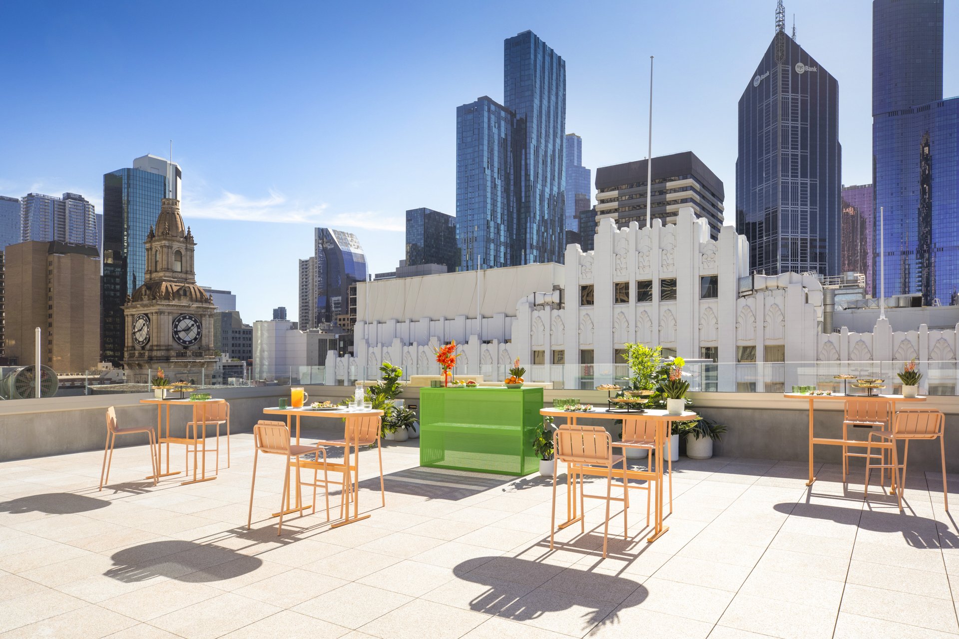 Outdoor terrace with tables and chairs, decorated with flowers and food, overlooking a skyline of modern skyscrapers and a historic clock tower, under a clear blue sky.