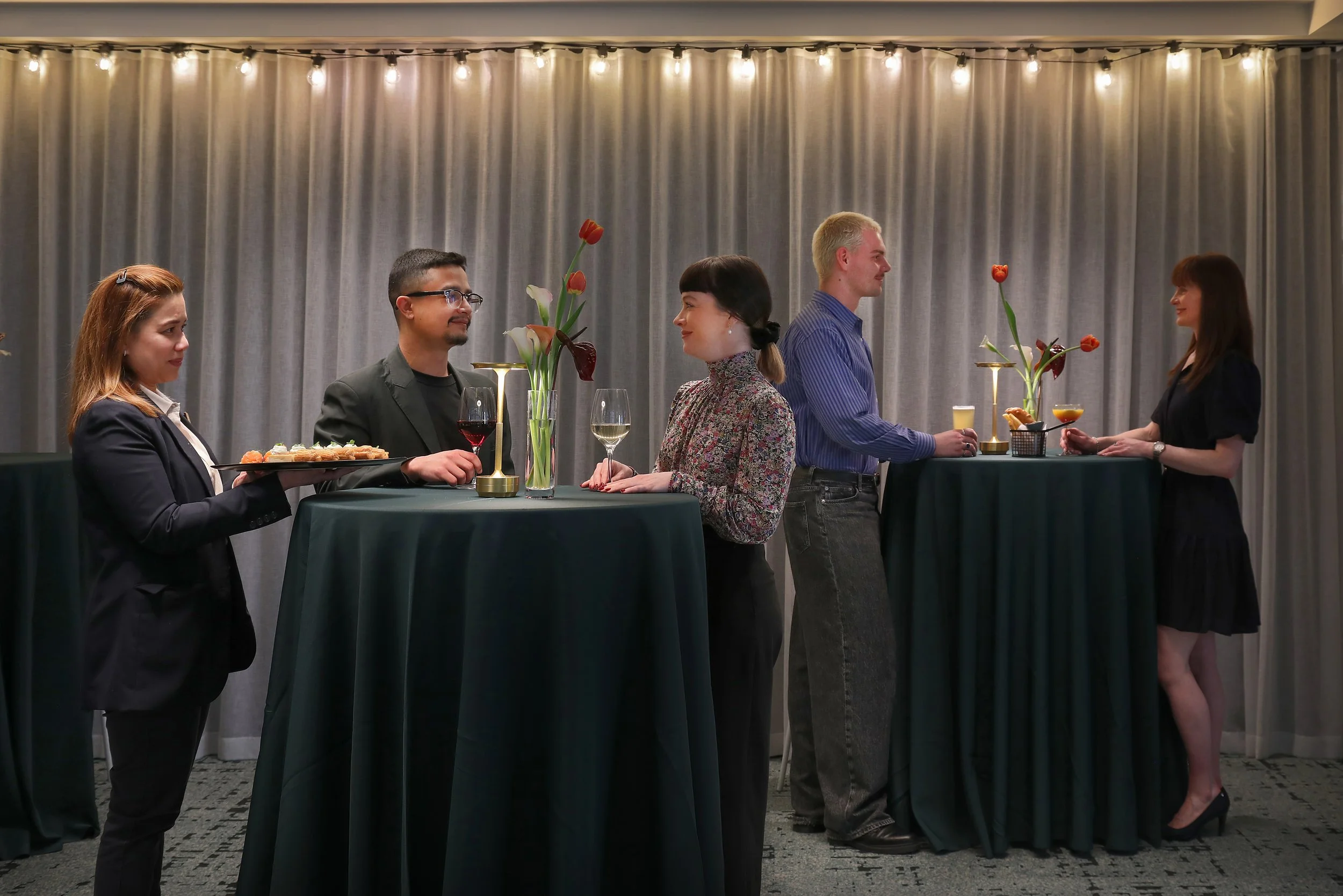 People at a social event around a tall high-top table with floral decor, drinks, and hors d'oeuvres, exchanging conversations in front of a curtain backdrop with string lights.
