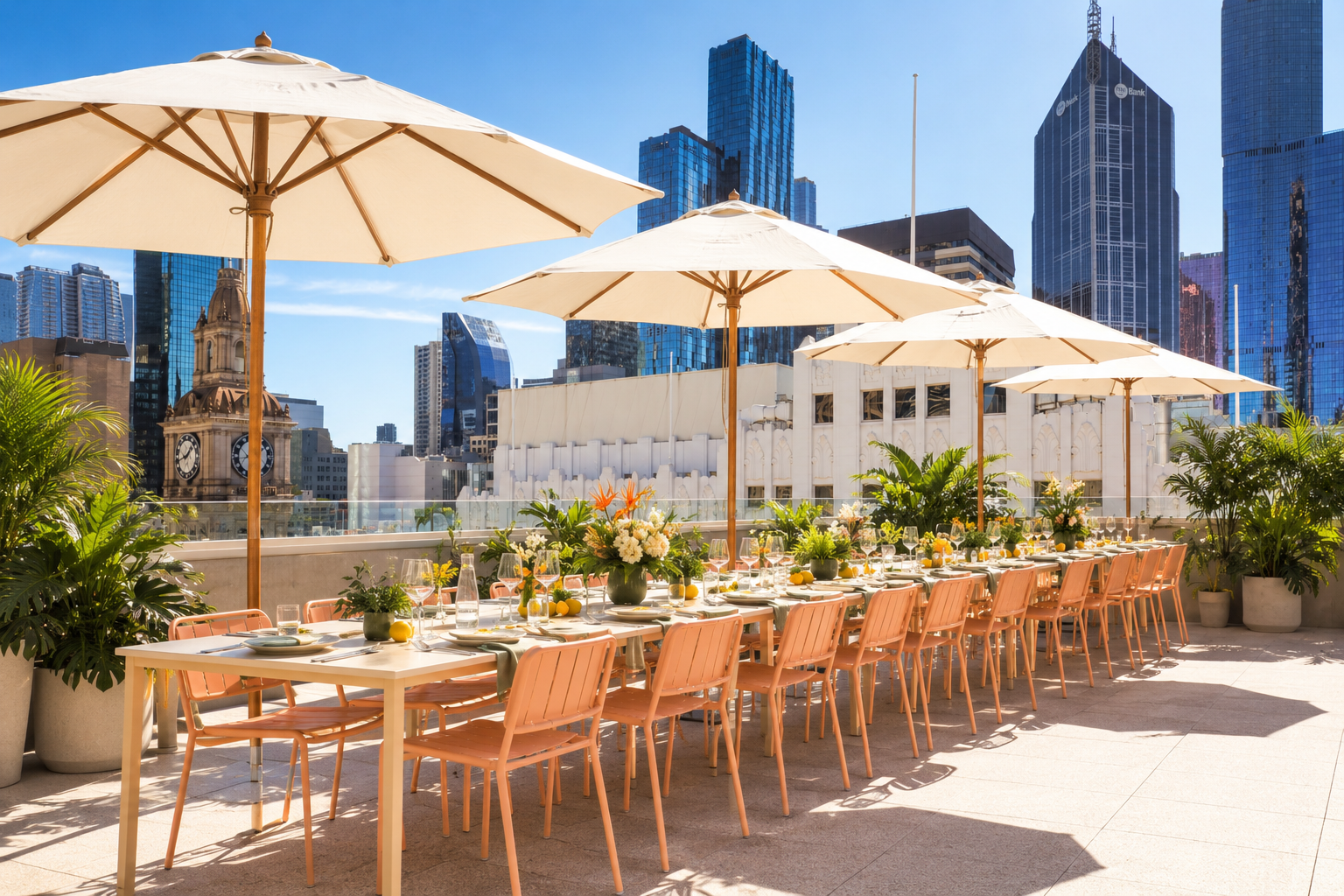 Outdoor rooftop dining setup with a long table, pink chairs, white umbrellas, potted plants, and city skyline in the background.