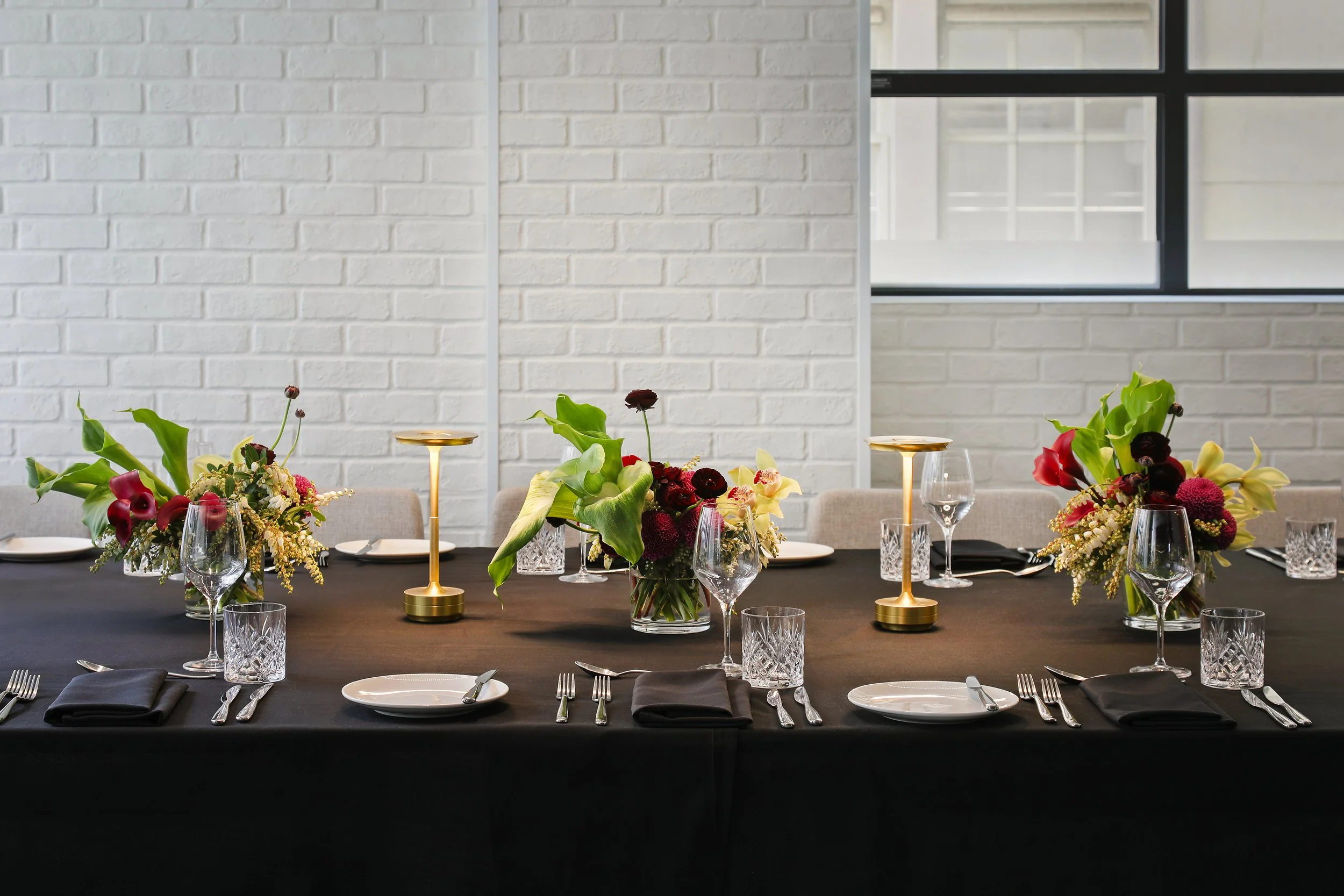 Table set for a formal event with floral centerpieces, black tablecloth, glassware, plates, black napkins, and silverware, against a white brick wall and window background.
