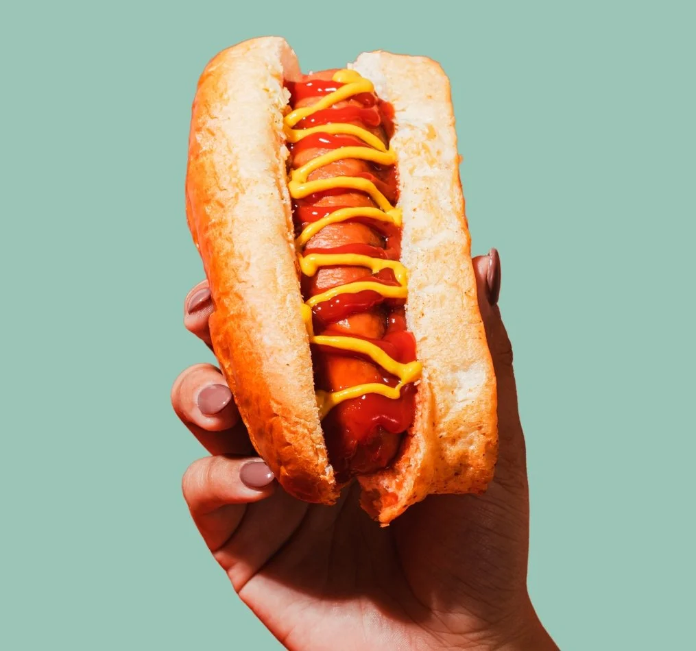 Close-up of a person holding a hot dog with ketchup and mustard on a light blue background.