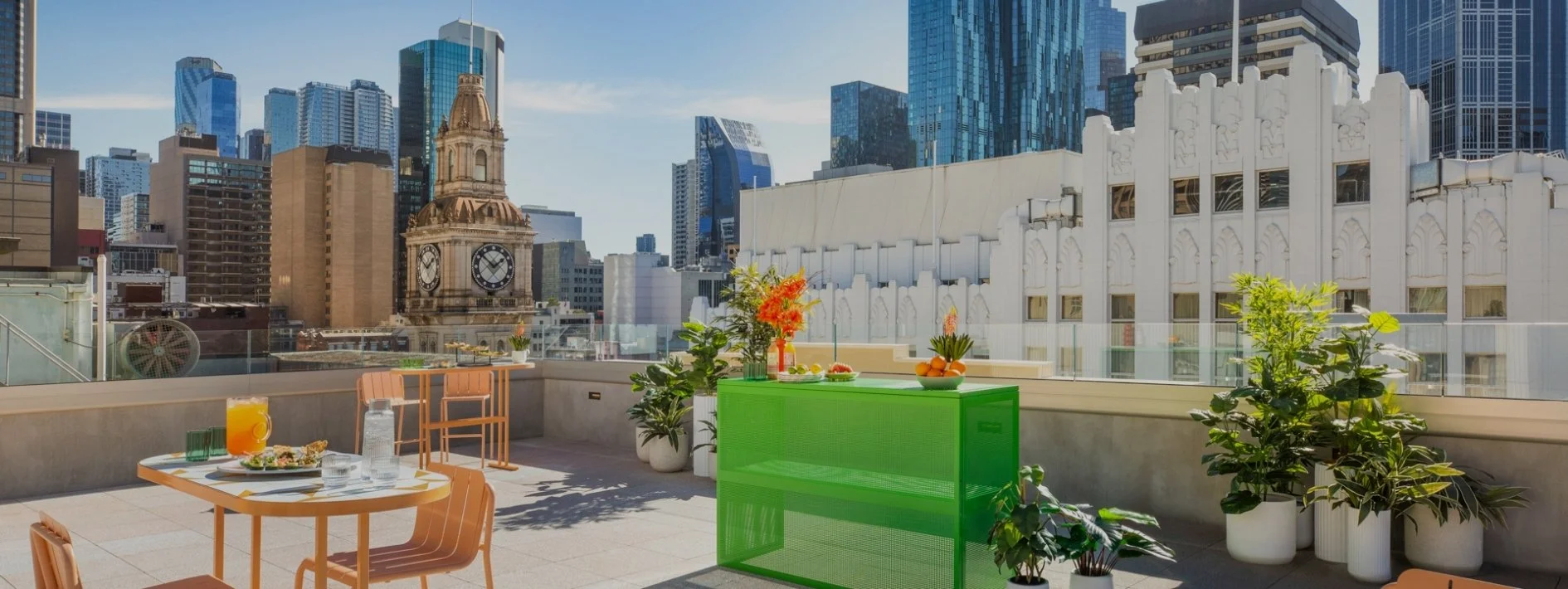 Rooftop terrace with tables, chairs, and plants overlooking a city skyline with tall modern buildings and a historic clock tower.