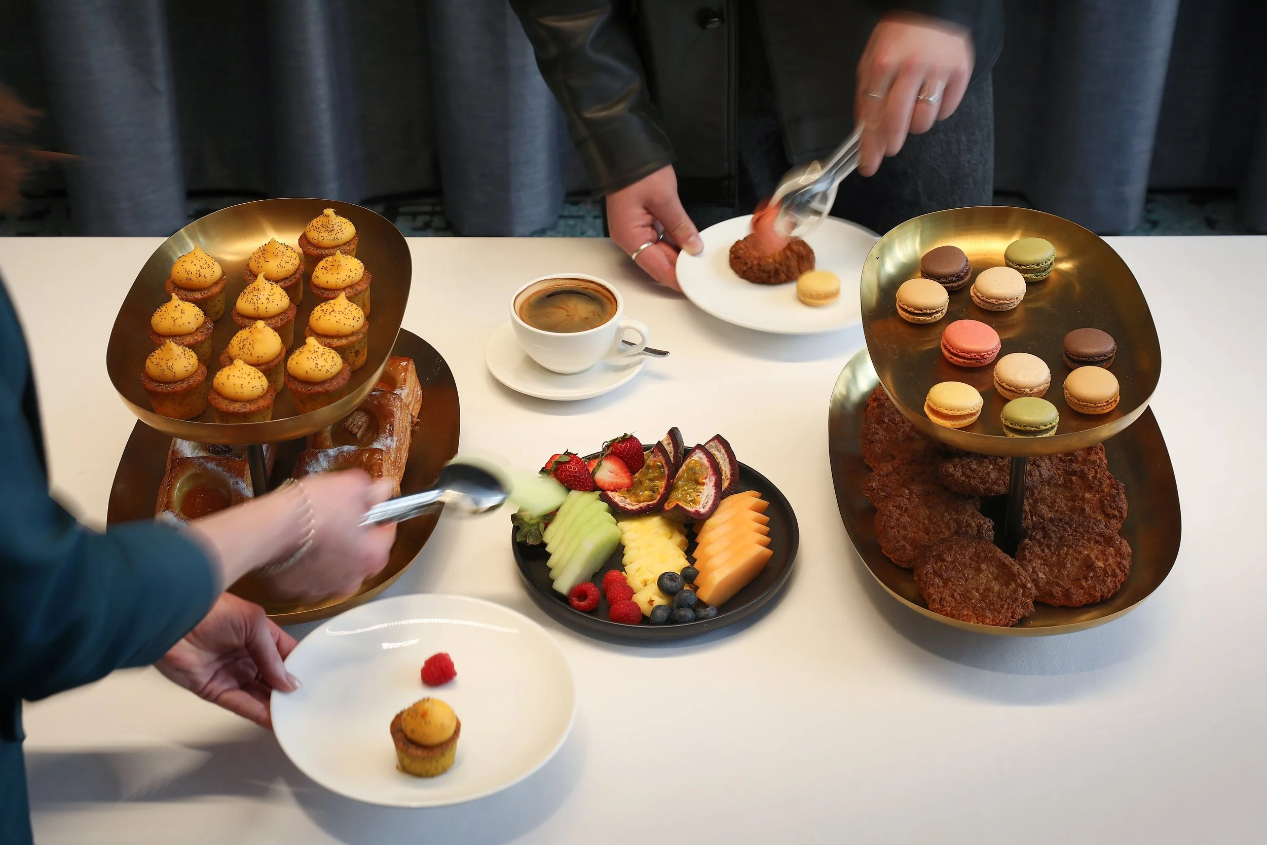 A person serving a small dessert cup with yellow frosting and black sprinkles onto a white plate at a dessert table that includes macarons, pastries, and fresh fruit.