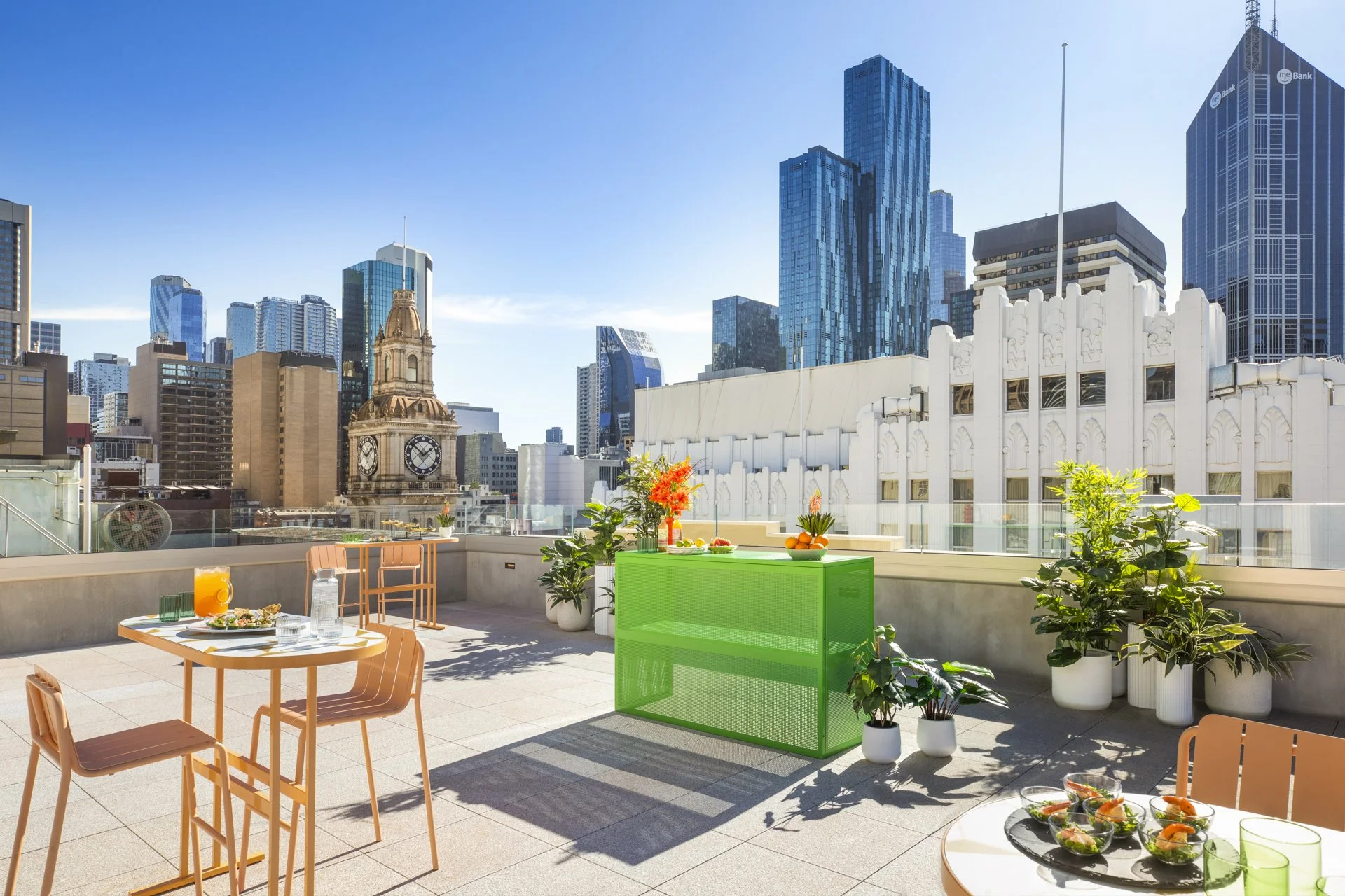 Rooftop terrace in downtown city with modern buildings and a historic clock tower in the background, decorated with potted plants, tables with food and drinks, and colorful outdoor furniture.