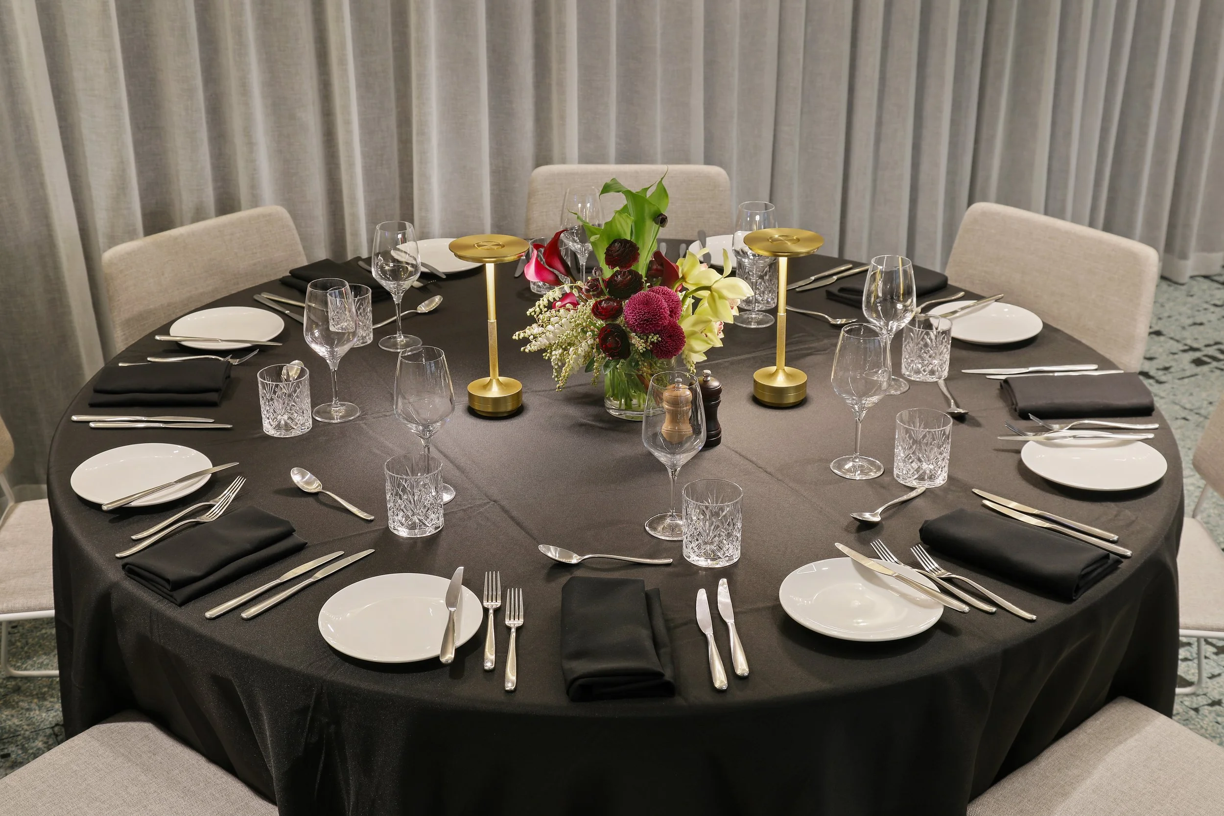A round banquet table with a black tablecloth, set with white plates, silver utensils, black napkins, wine glasses, and water glasses. In the center, there is a floral arrangement with pink, purple, white, and green flowers, and two gold candleholder