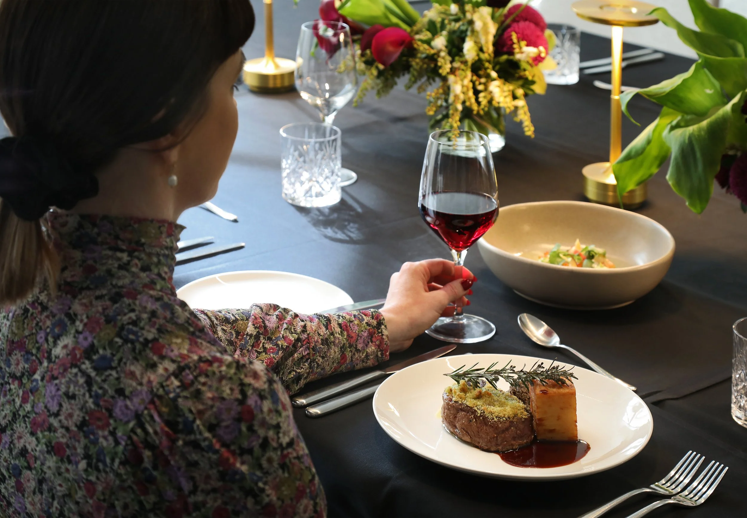 A woman in a floral dress sits at a dining table, holding a glass of red wine. The table is set with a white plate containing a piece of beef Wellington, and other elegant tableware. In the background, there is a large floral centerpiece with pink, y