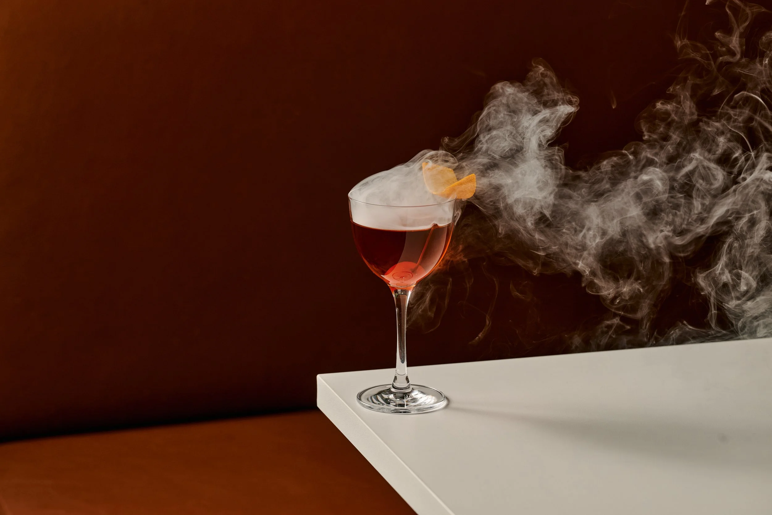 A wine glass with red liquid on a white surface, with smoke or vapor rising and swirling around it against a dark background.