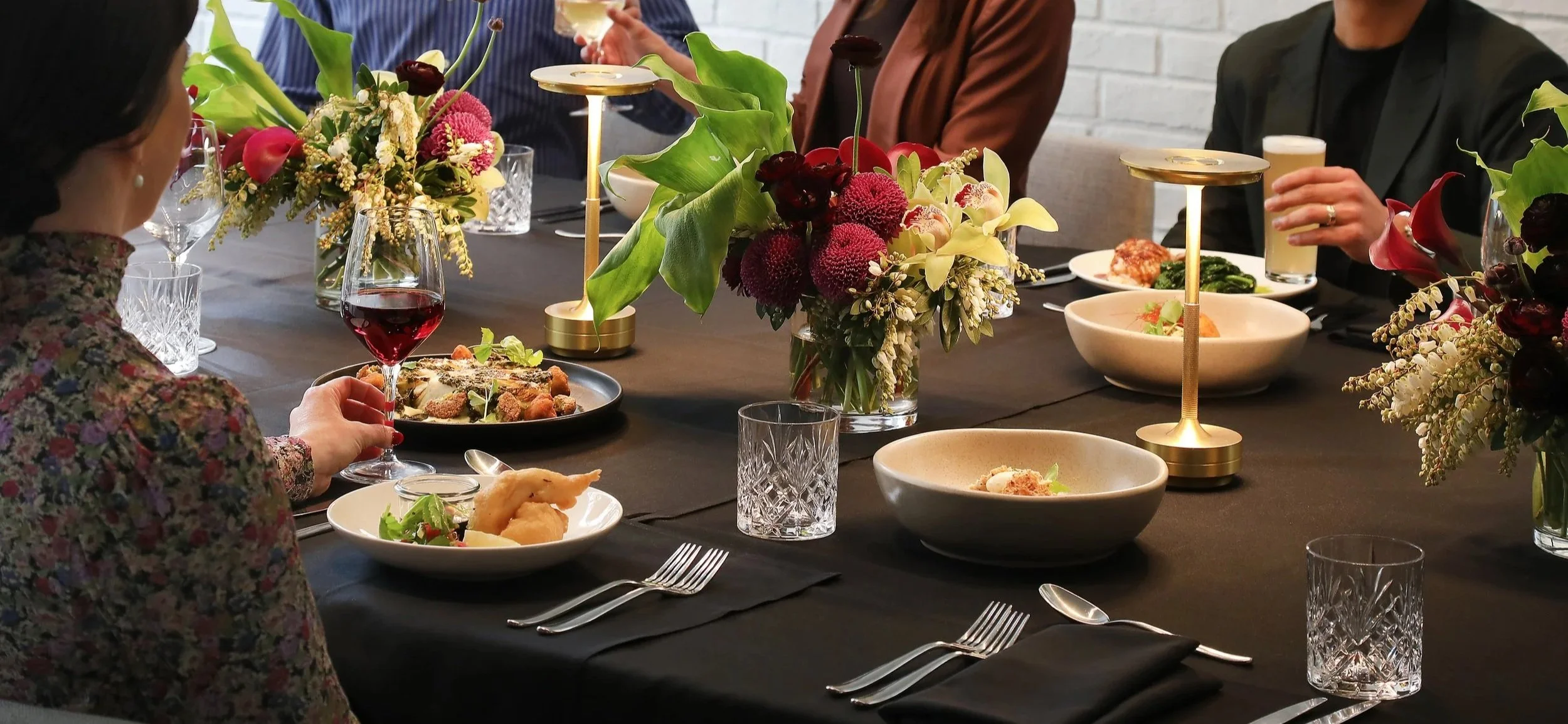 People sitting at a dinner table with floral centerpieces, wine glasses, and dishes of food, including fried items and salads, in a well-lit room with a brick wall in the background.