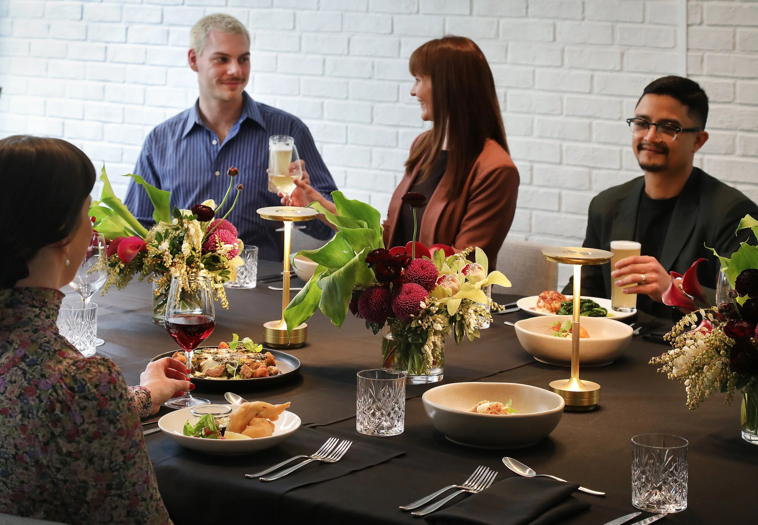 People at a dinner party with food and flowers on a black table against a white brick wall.