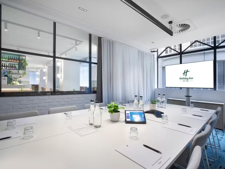 A modern conference room with a large white table, water bottles, glasses, and notepads, overlooking a cityscape through large windows, with a flat-screen monitor displaying the Holiday Inn logo.