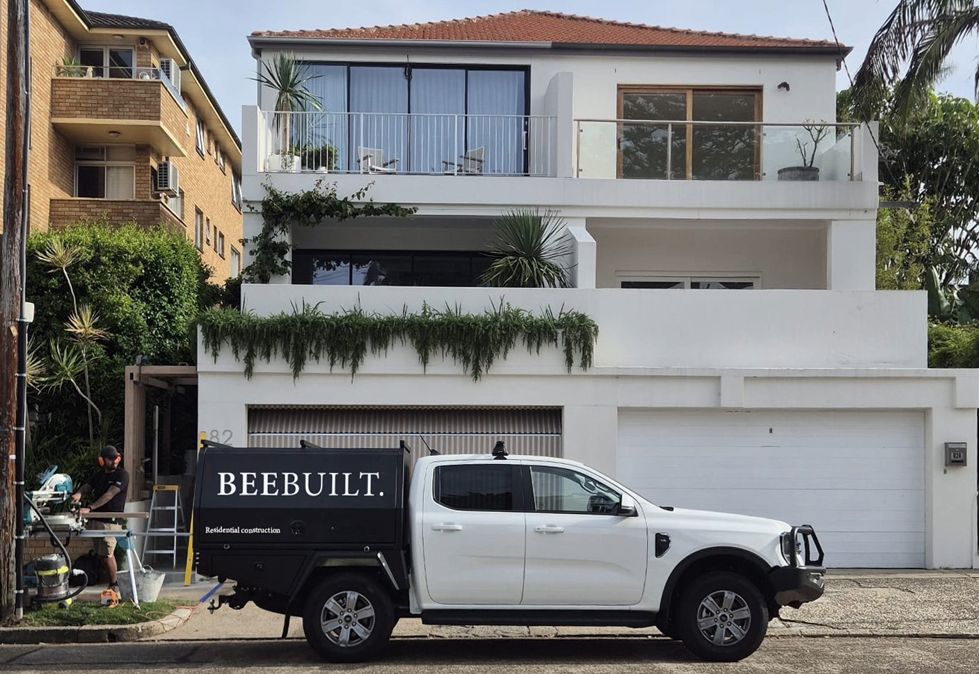 White construction truck parked in front of a recently renovated modern white residence with two balconies and a worker nearby. The truck has a black container with the brand name 'BEEBUILT.' in white lettering.