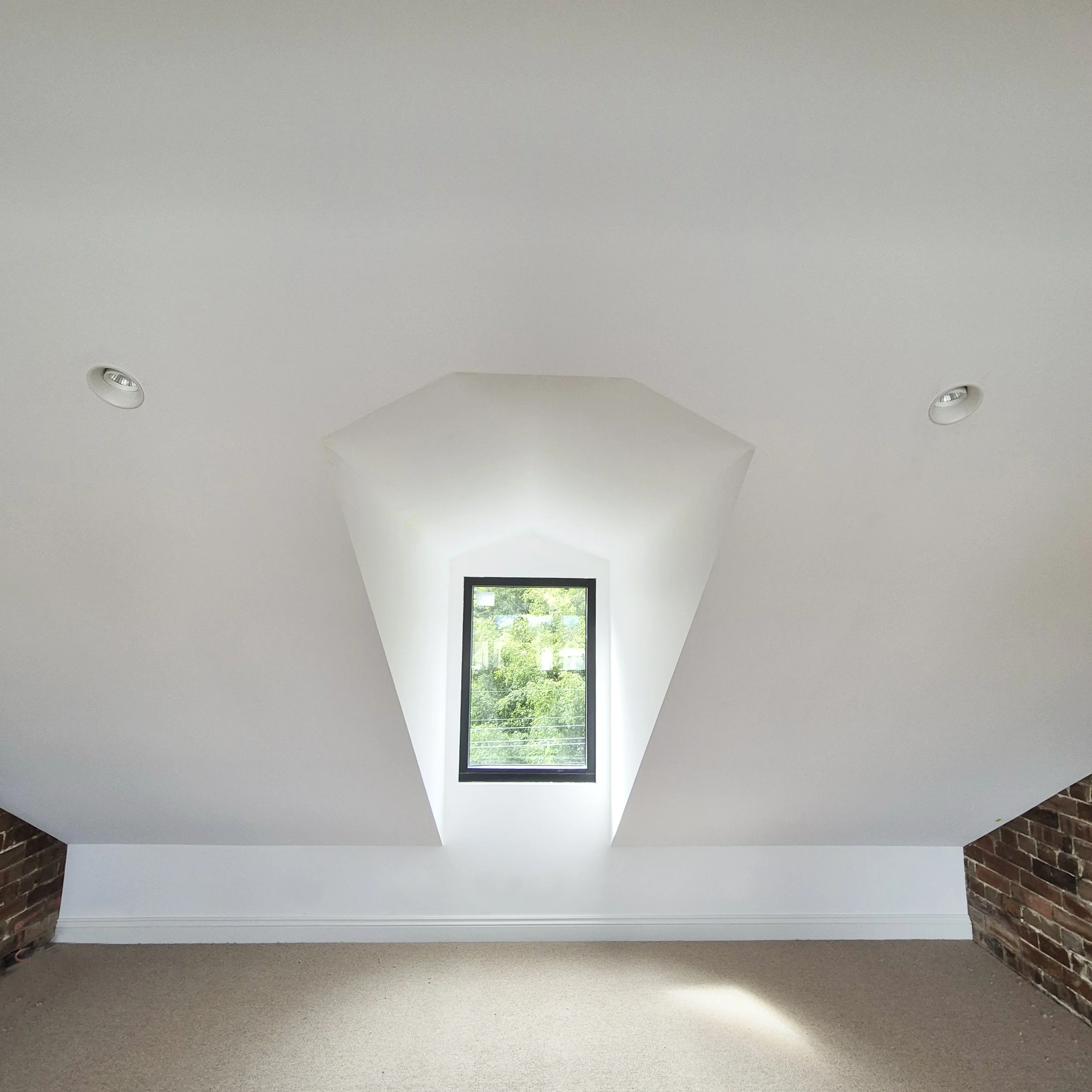Photo of a white ceiling with two recessed lights and a rectangular window letting in natural light, showing trees outside.