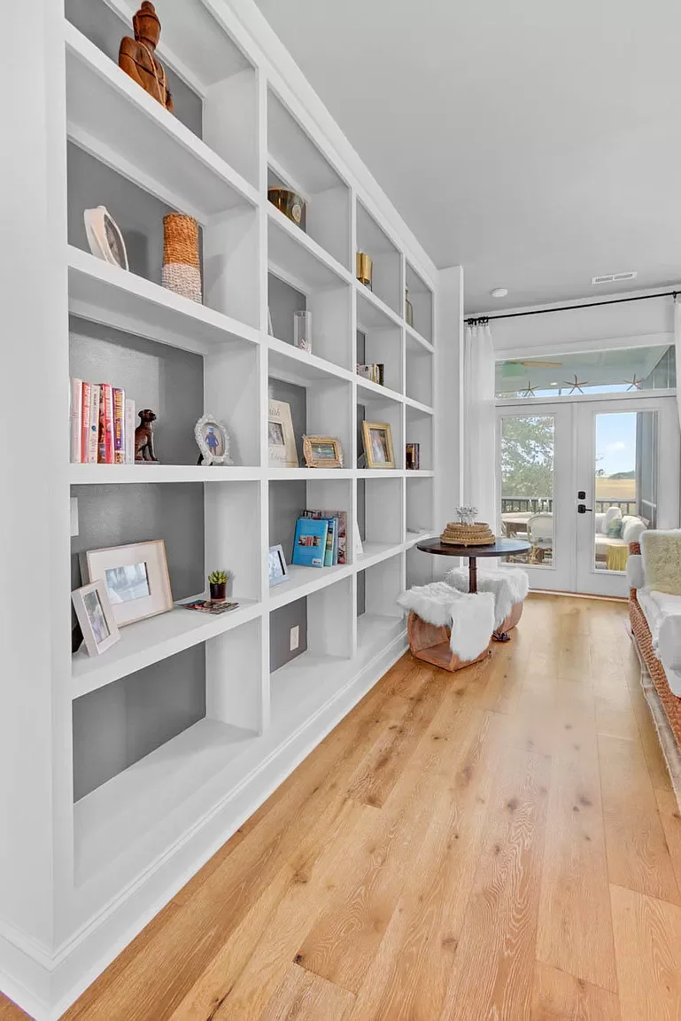 Living room with white built-in wall shelves, decorated with framed photos and small decorative items. Hardwood floor, a small round table with a fluffy white cover, and a glass door leading to an outdoor patio with scenery.