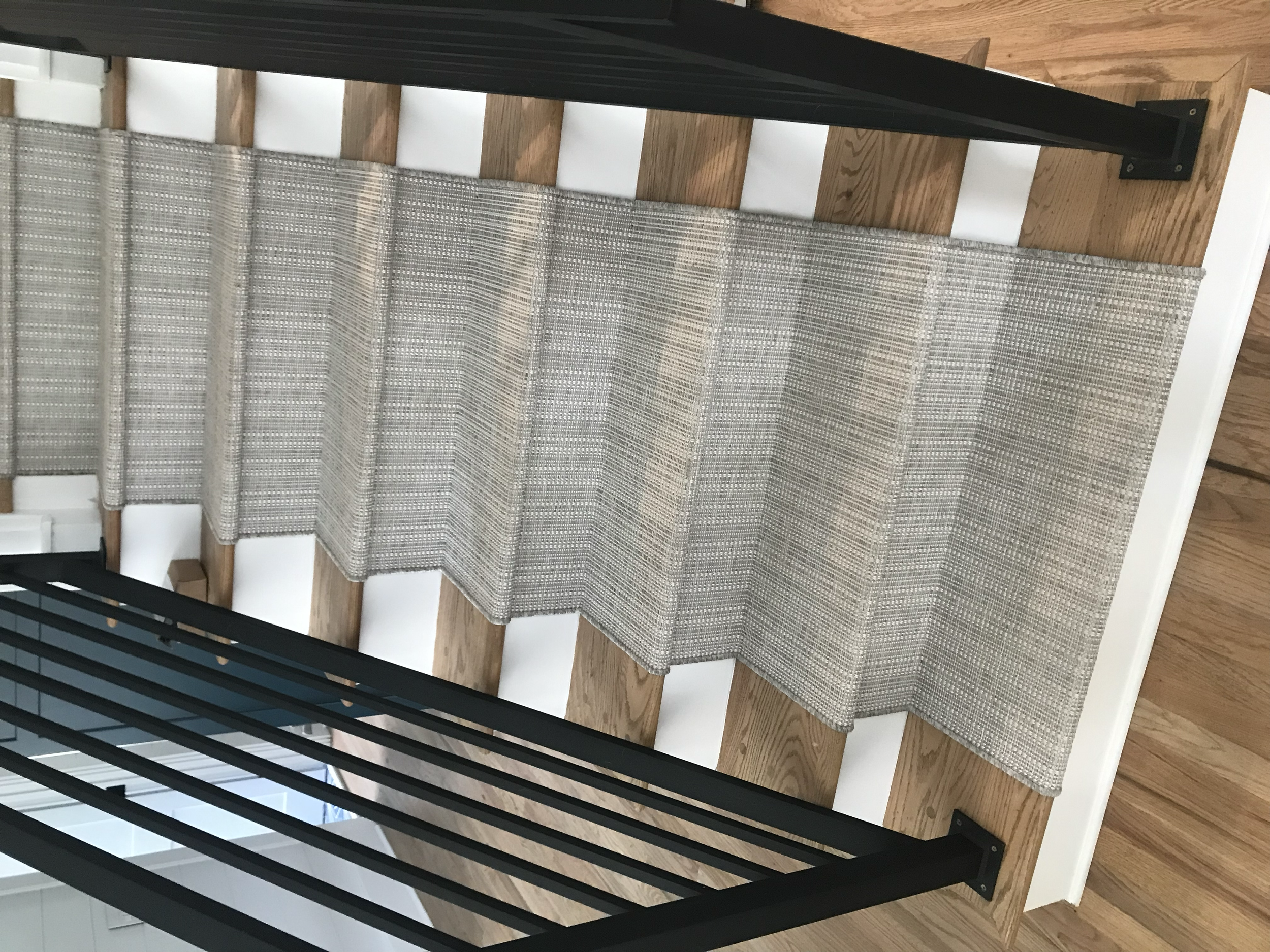 A staircase with a wood railing and carpeted steps. The staircase has a modern design with black metal balusters, and the steps are covered in a woven patterned carpet runner. The floor is wooden, and part of a white wall is visible at the bottom.